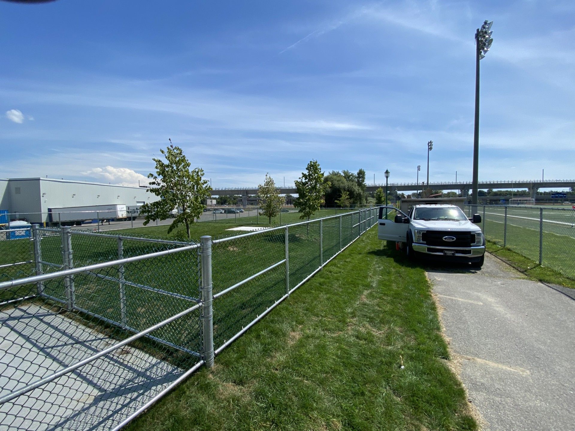 Chain link fence bordering a grass area and a paved path. A white truck parked alongside. Blue sky.