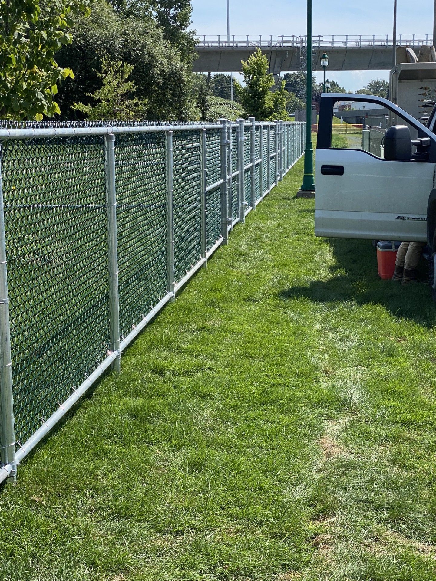 Chain link fence with green privacy slats bordering green grass, a bridge, and a white truck.