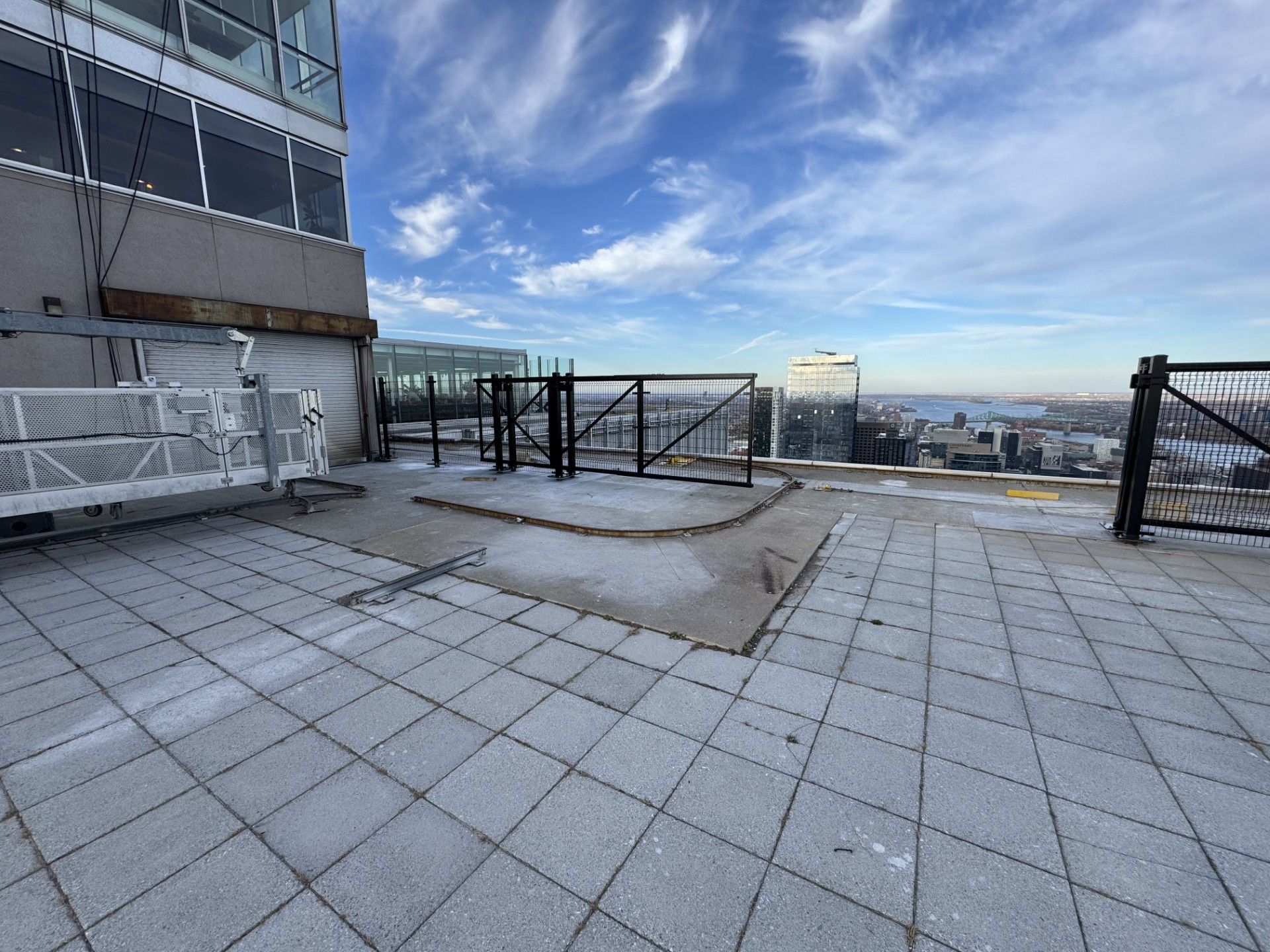 Rooftop patio with light-colored square tiles, metal fencing, and views of cityscape under a cloudy blue sky.