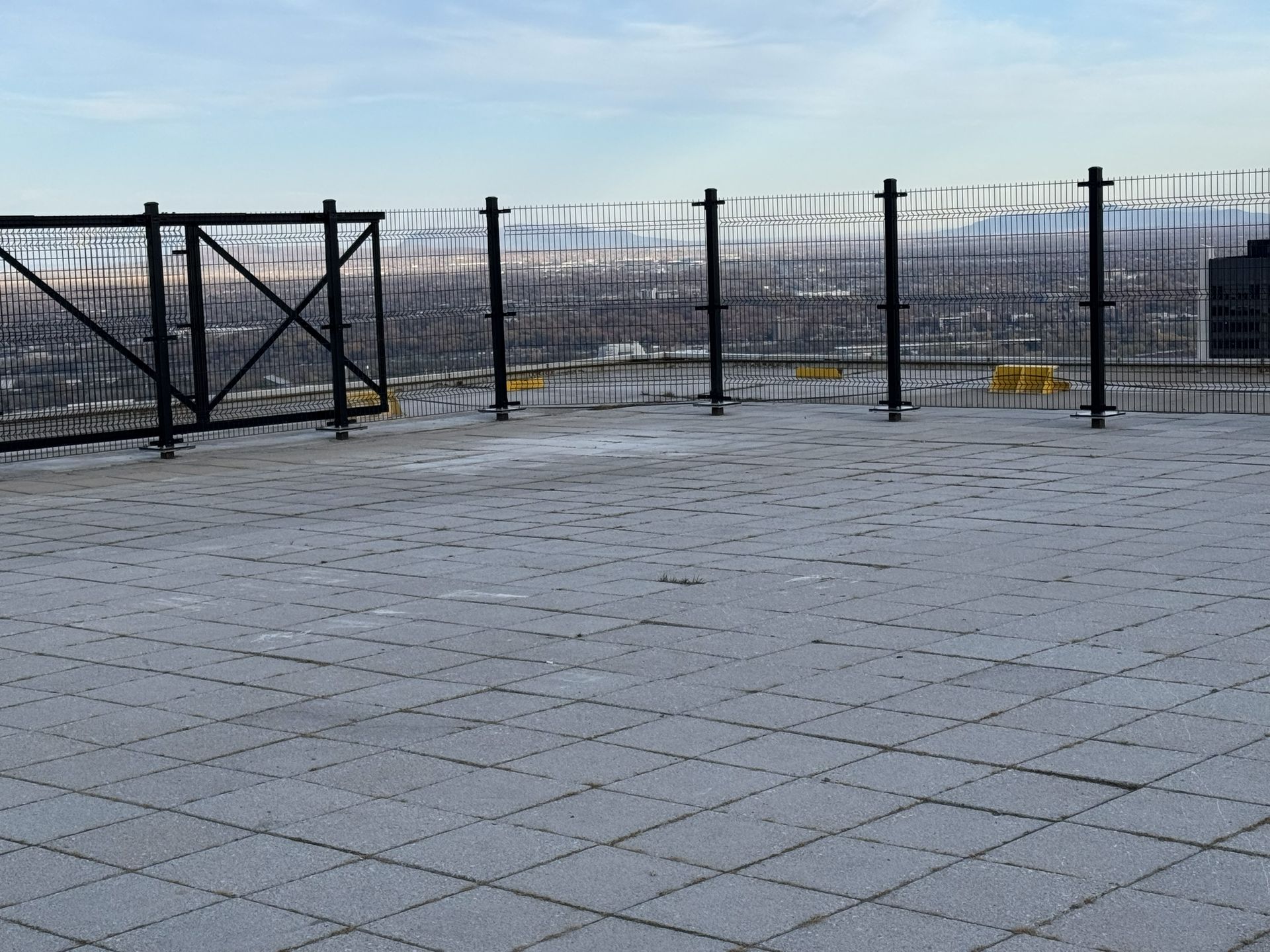 Rooftop patio with light-colored tiles, black fencing, and a distant city view under a hazy sky.
