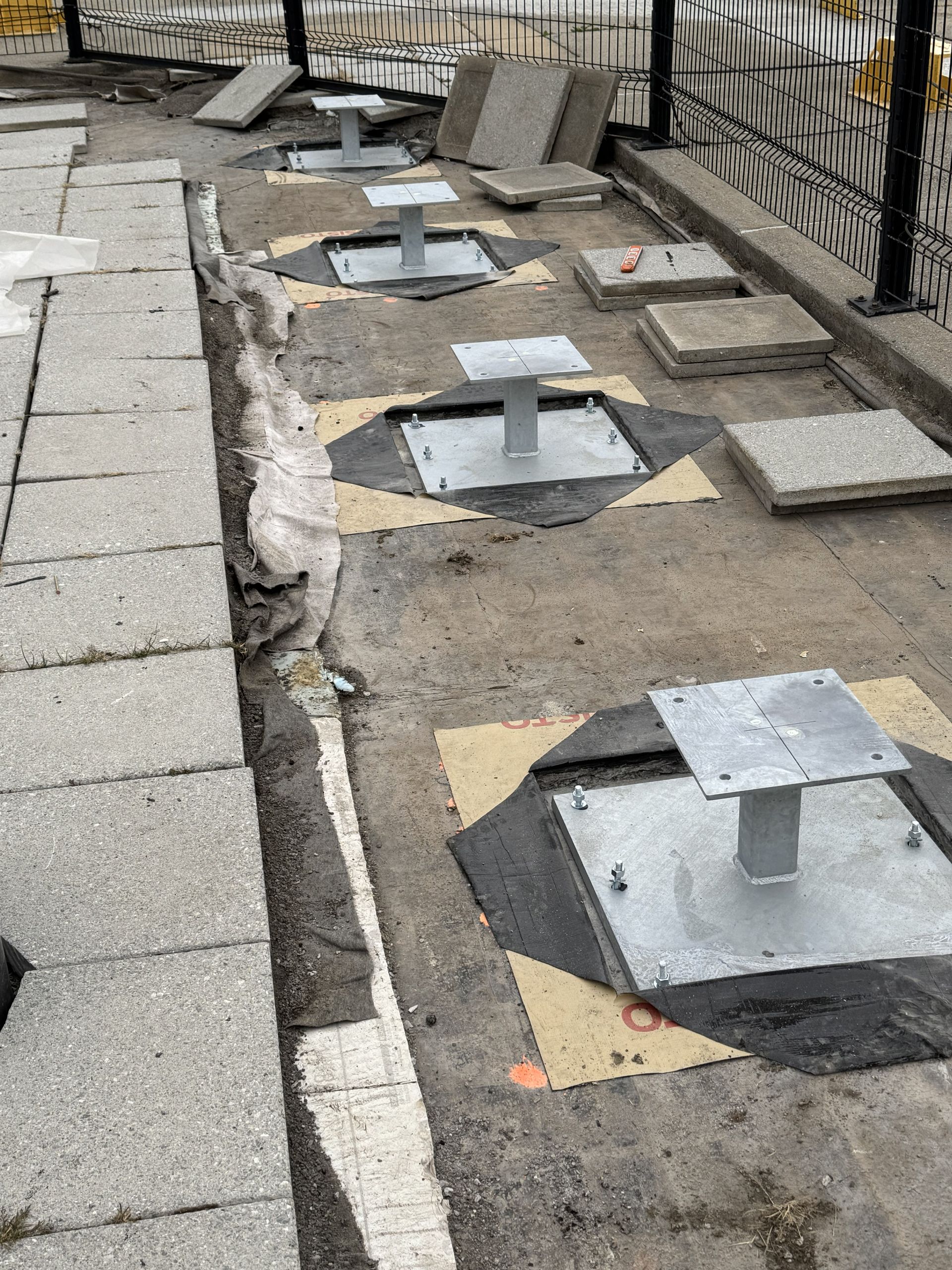 Metal posts mounted in the ground, with surrounding construction, near a sidewalk.