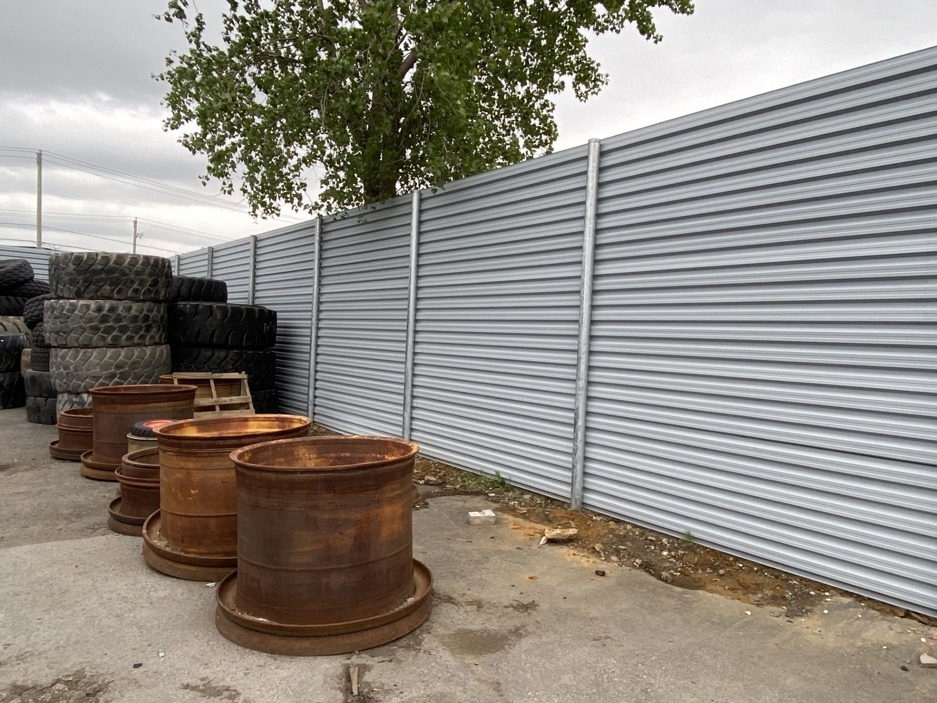 Stack of tires and rusty metal objects in front of a corrugated metal fence, with a tree and cloudy sky visible.