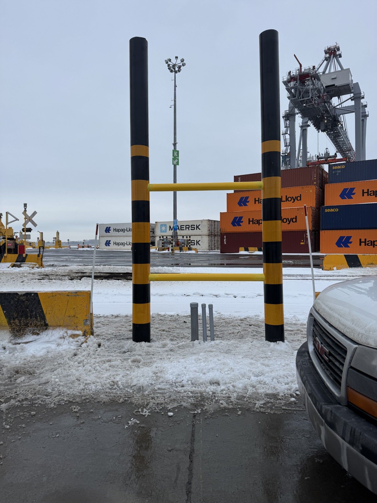 Black and yellow striped gate at a port, snow on the ground, shipping containers and crane in the background.