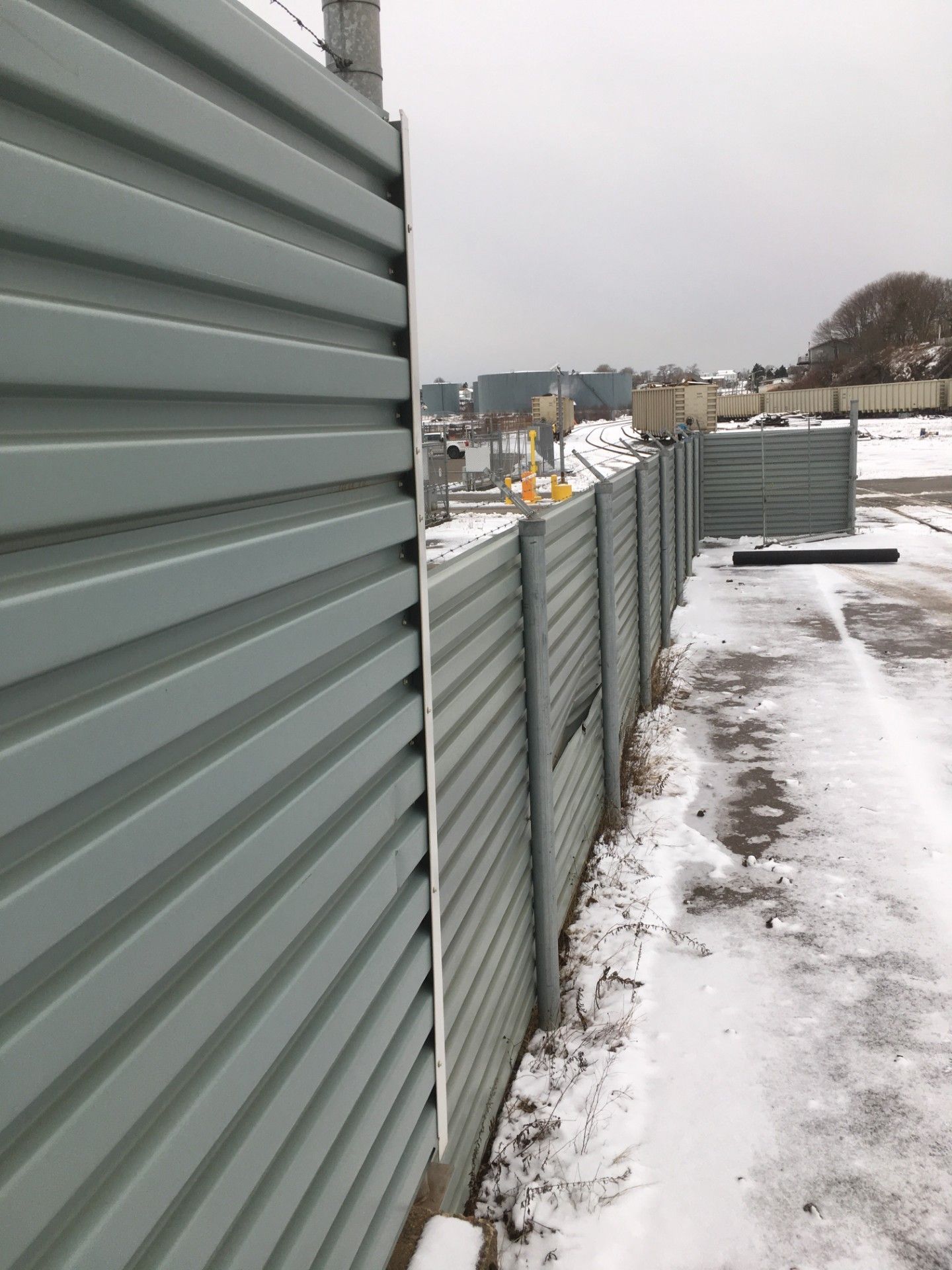 Gray corrugated metal fence, snow-covered ground, overcast day. Industrial area visible beyond.