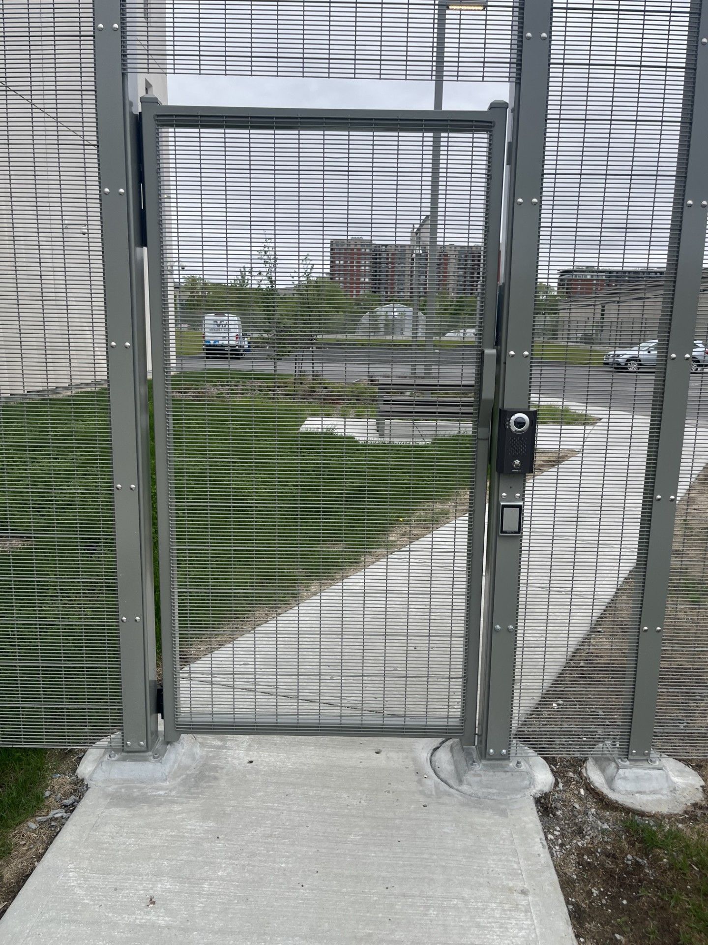 Metal gate in a chain-link fence on a concrete base, partially open. Cars and a building are visible in the background.