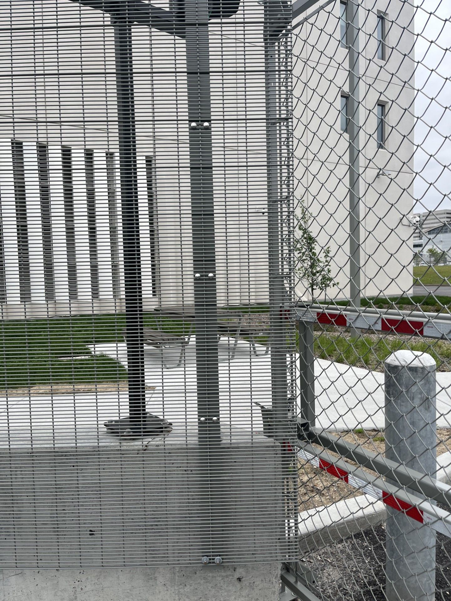 Metal security gate with chain-link fence, concrete base, and buildings in background.