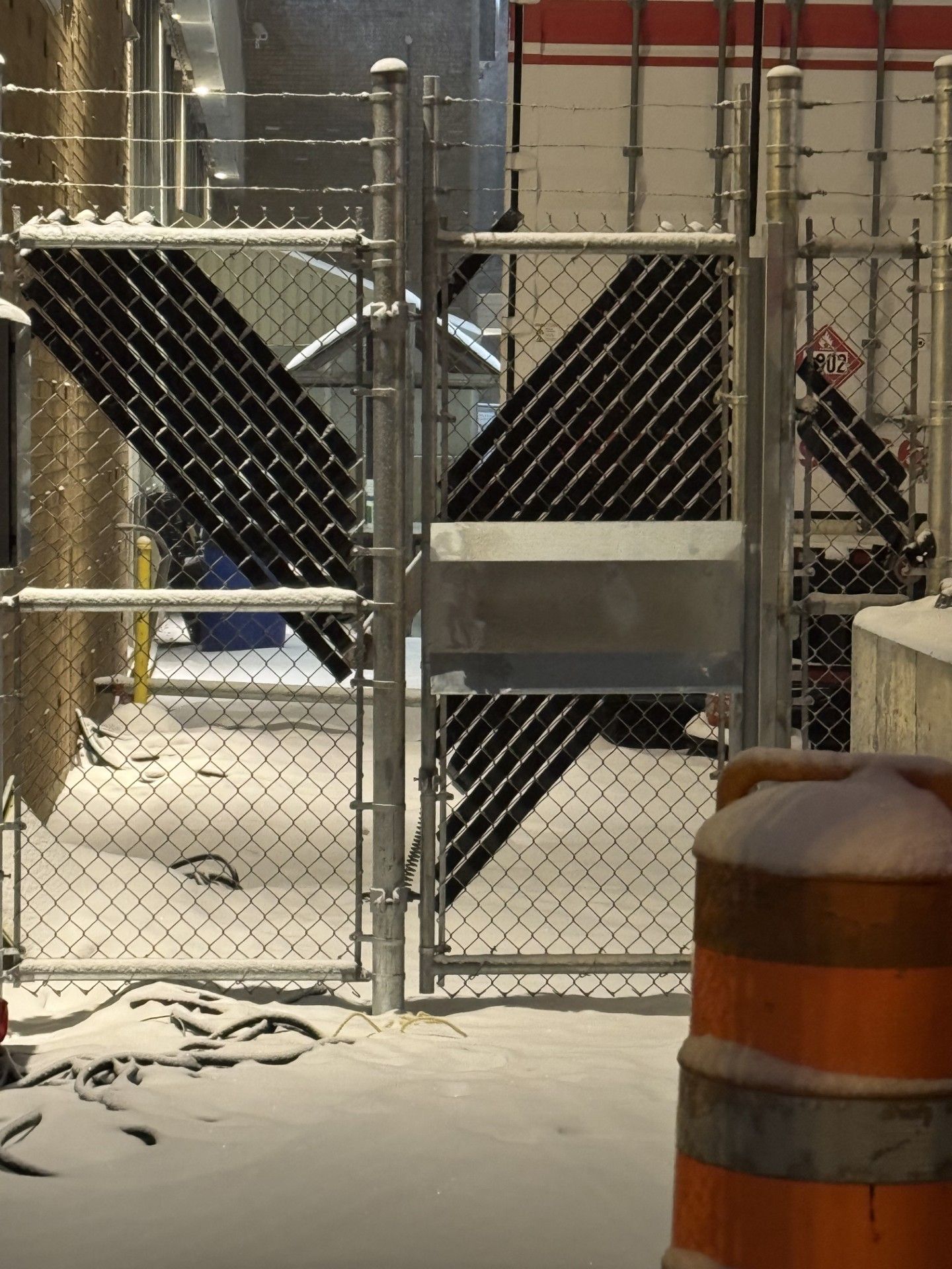 Snowy alleyway with chain-link gate. Black diagonal stripes on the gate, orange traffic cone in foreground.