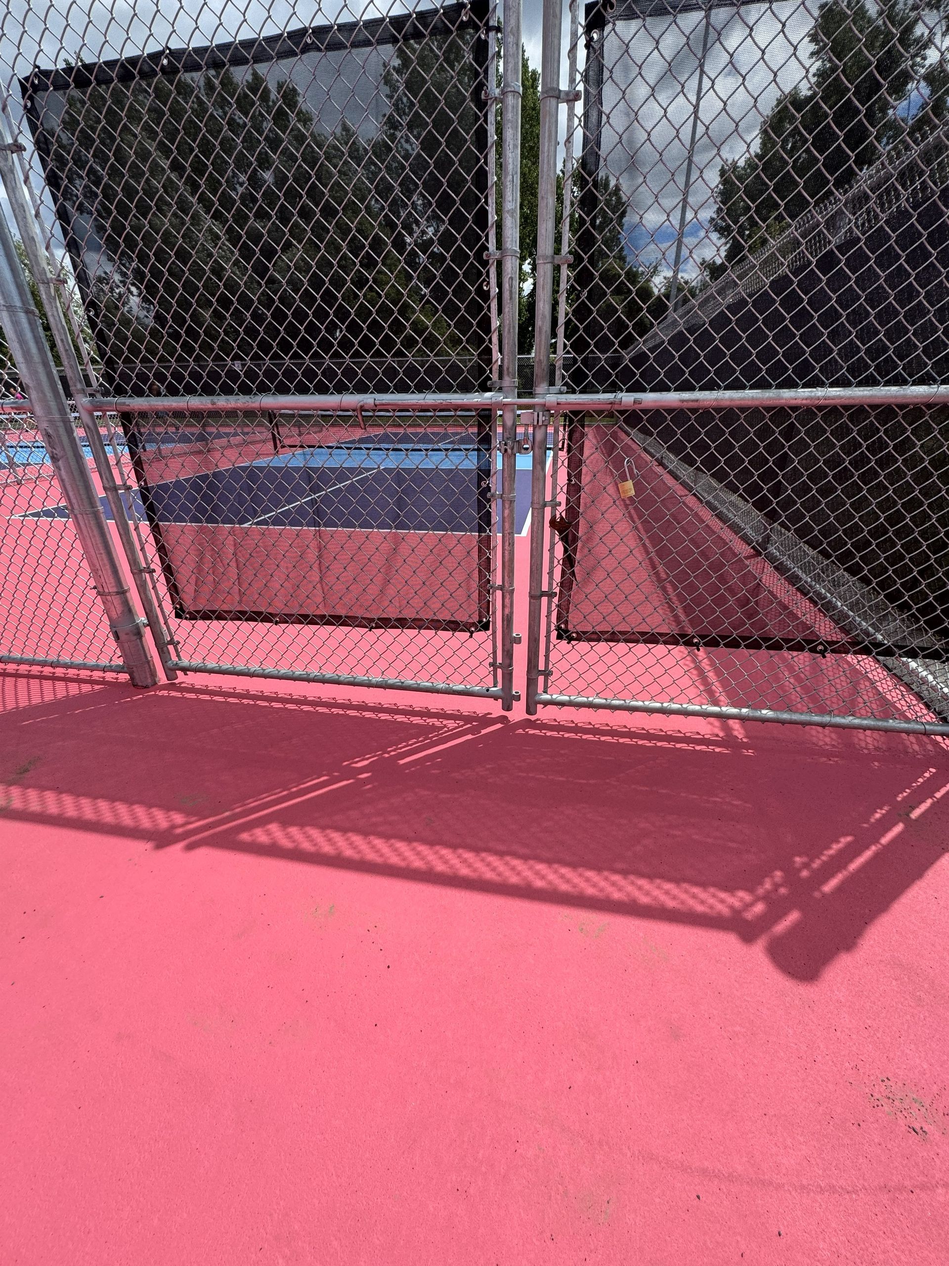 A pink painted tennis court, seen through a chain-link fence and gate, with shade cloth.