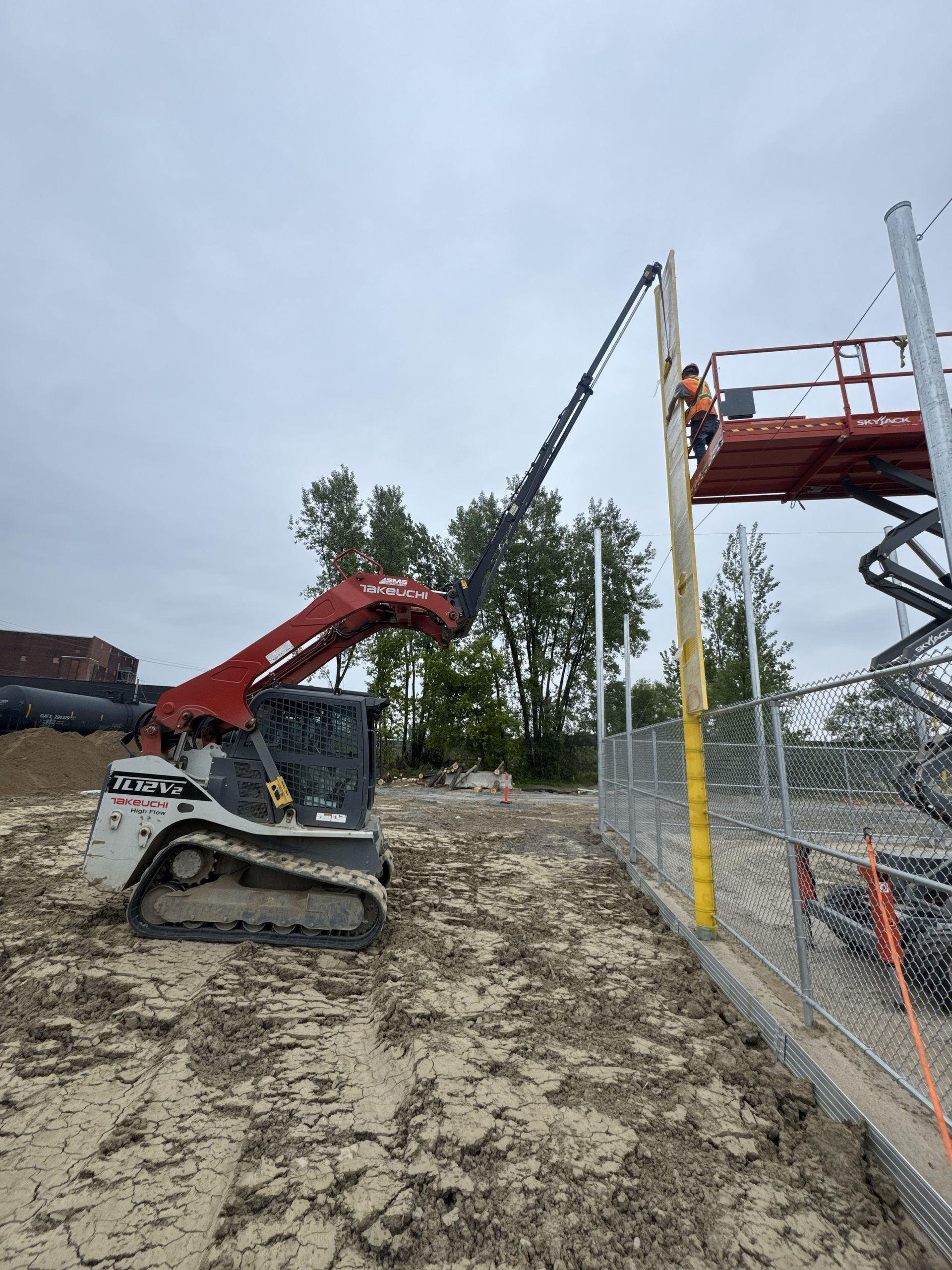 A skid steer and lift work together to install a post near a chain-link fence on a muddy construction site.