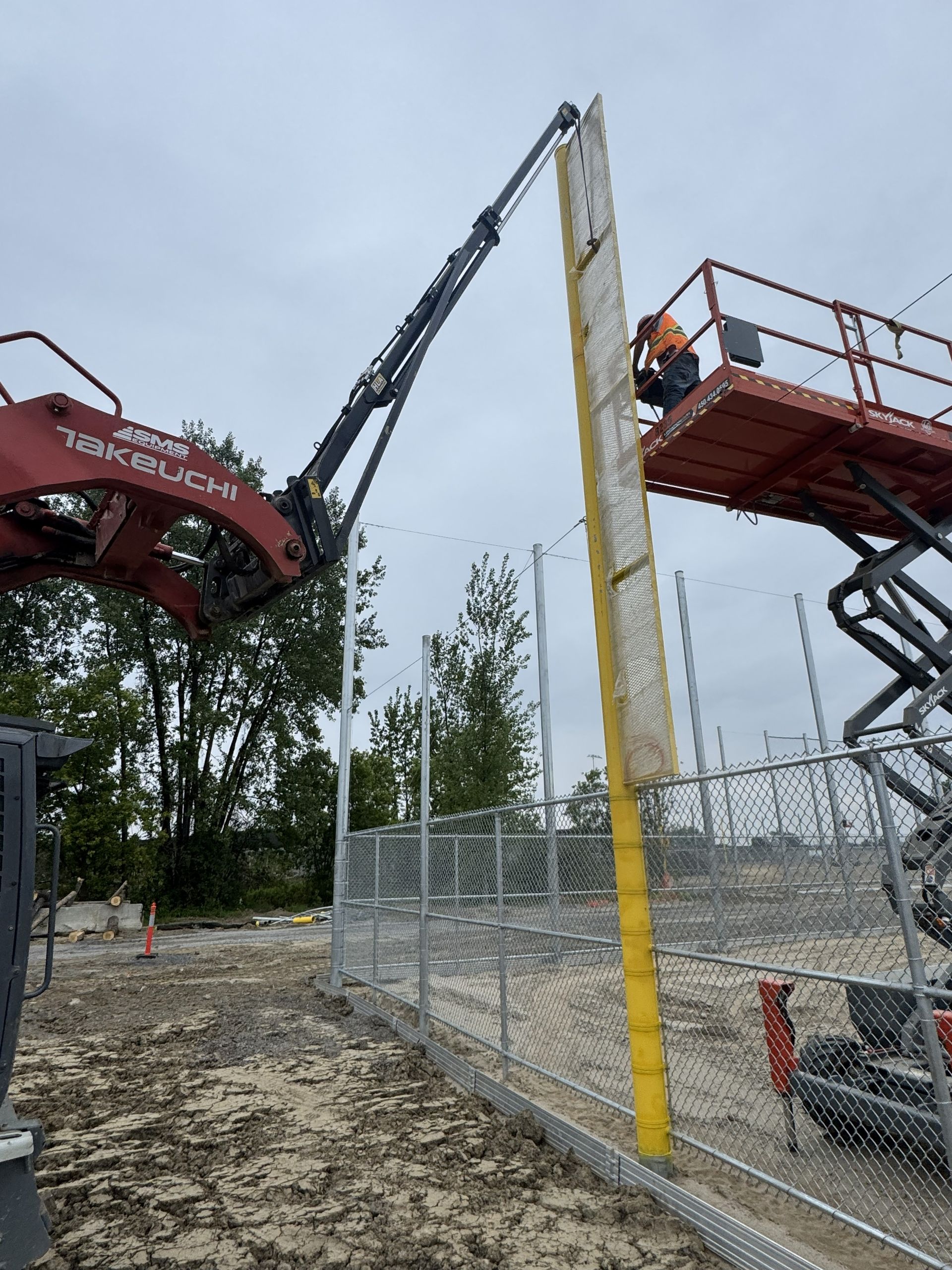 Des ouvriers du bâtiment installent un poteau de clôture à l'aide d'une grue et d'une nacelle élévatrice sur un chantier.