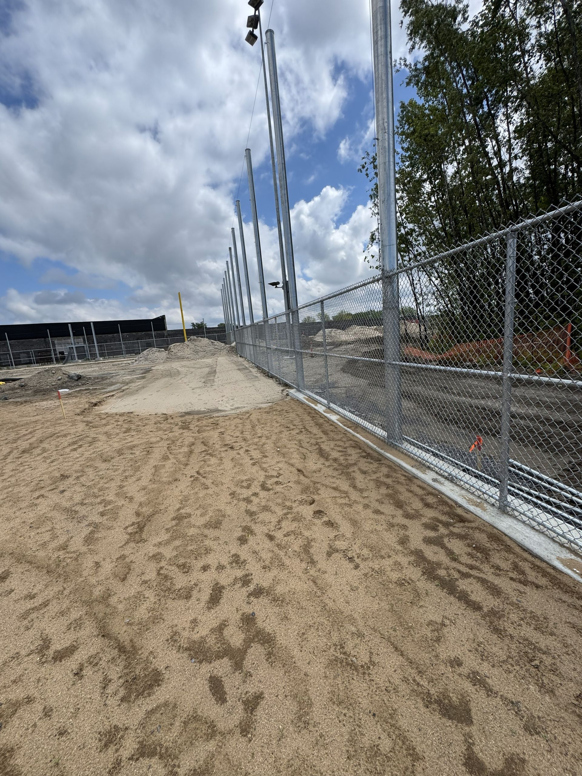 Gravel path next to a chain-link fence, under a cloudy sky. Metal light poles stand along the fence.