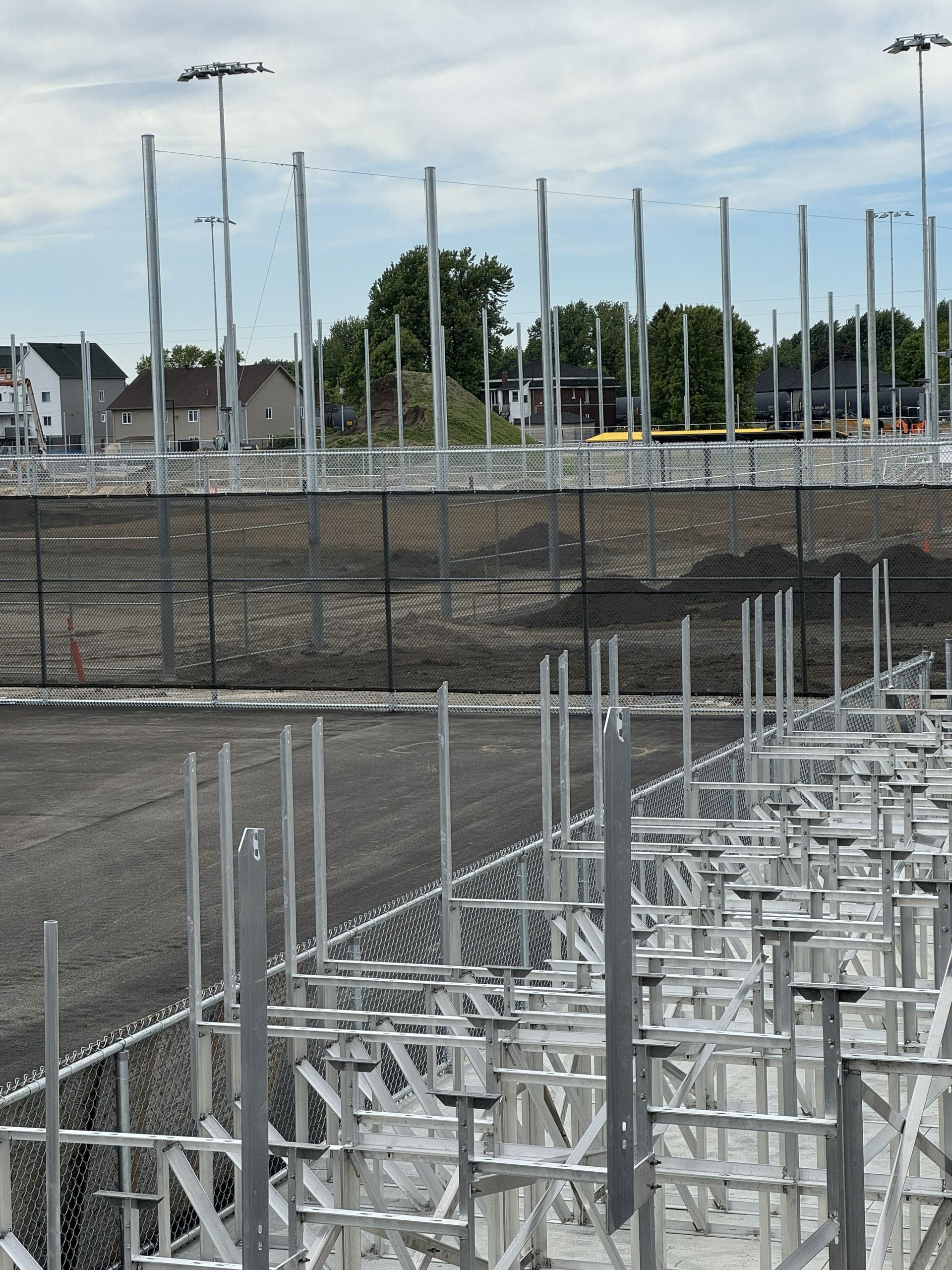 Metal framework construction, likely for a sports facility, with a fence in the foreground and a residential area in the background.