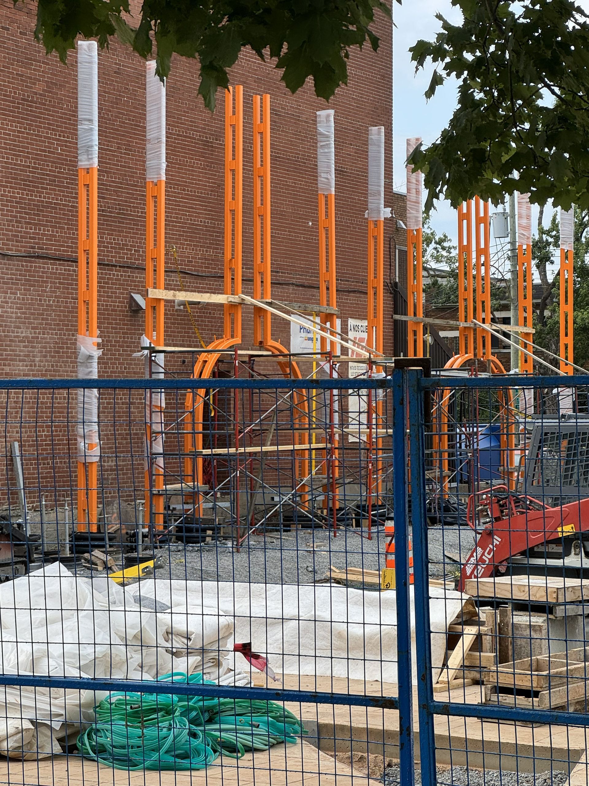 Construction site with orange scaffolding against a brick building, behind a blue safety fence.