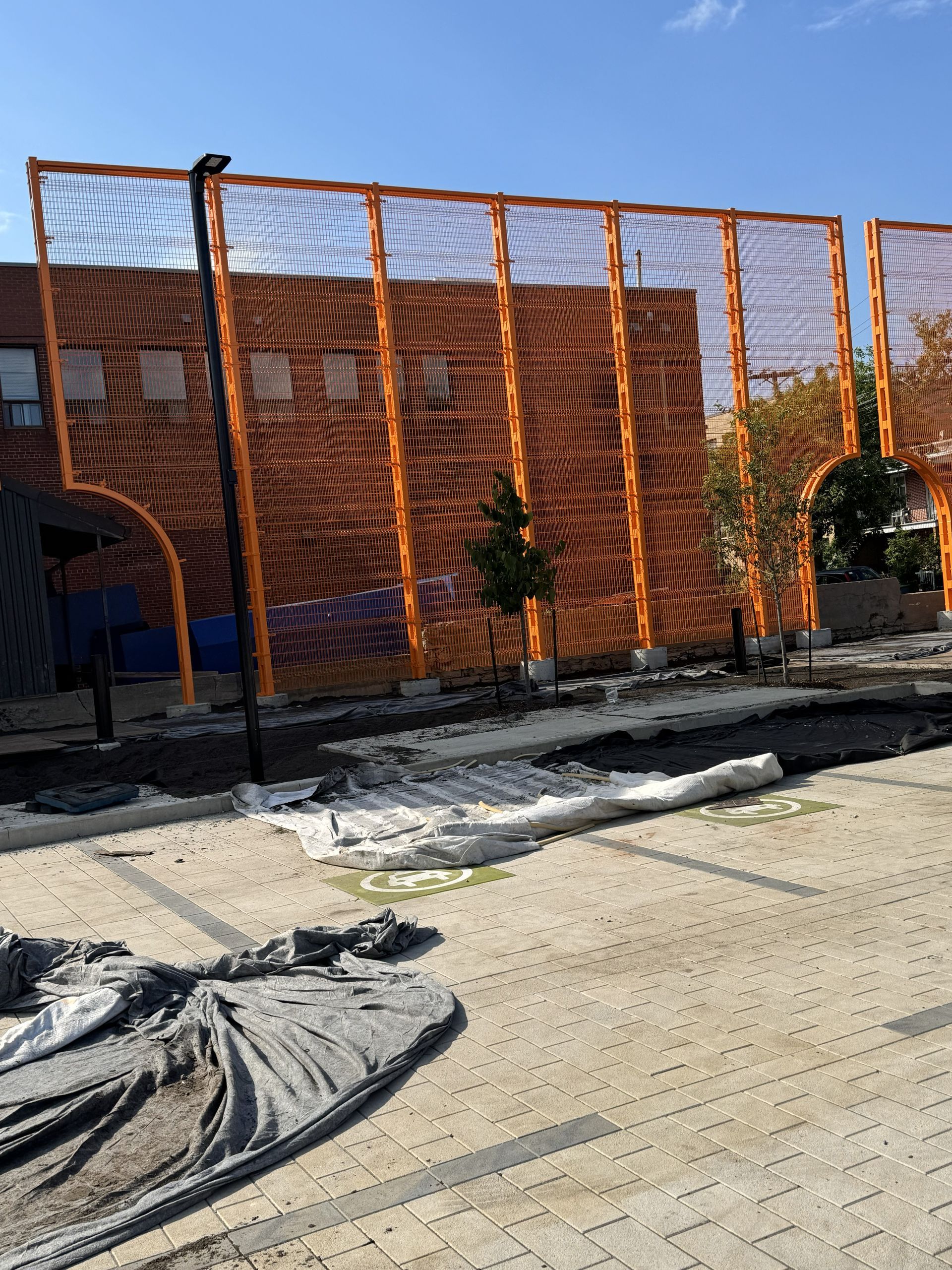 Orange mesh fence along a paved area with a small tree in the center. A building is behind the fence.