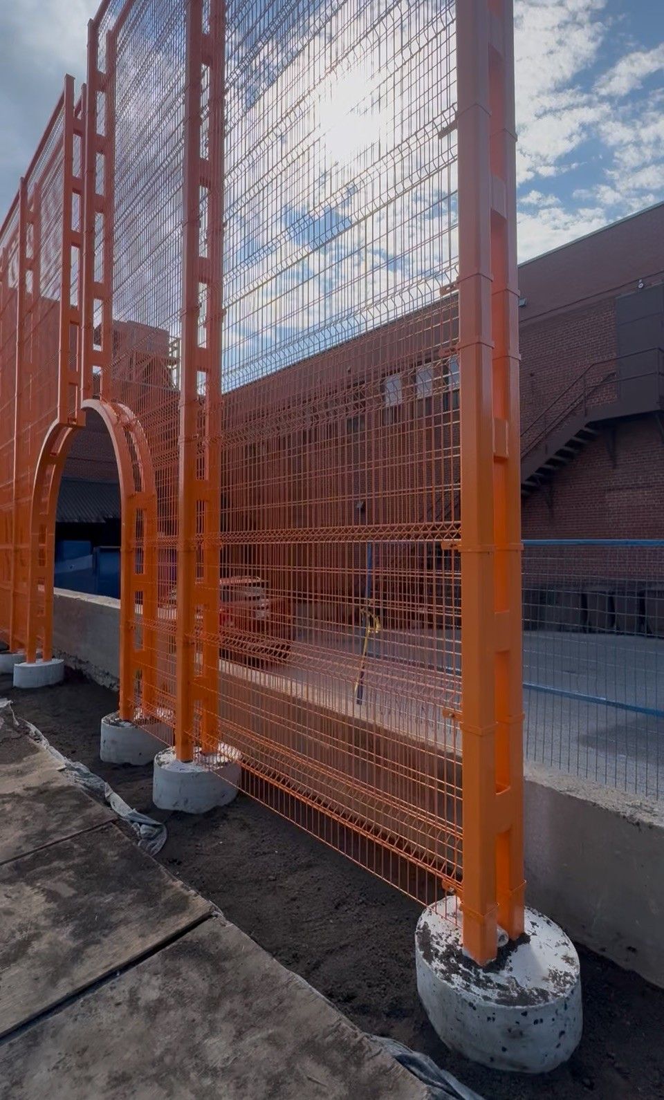 Orange metal fence with concrete footings against a building with a blue sky.