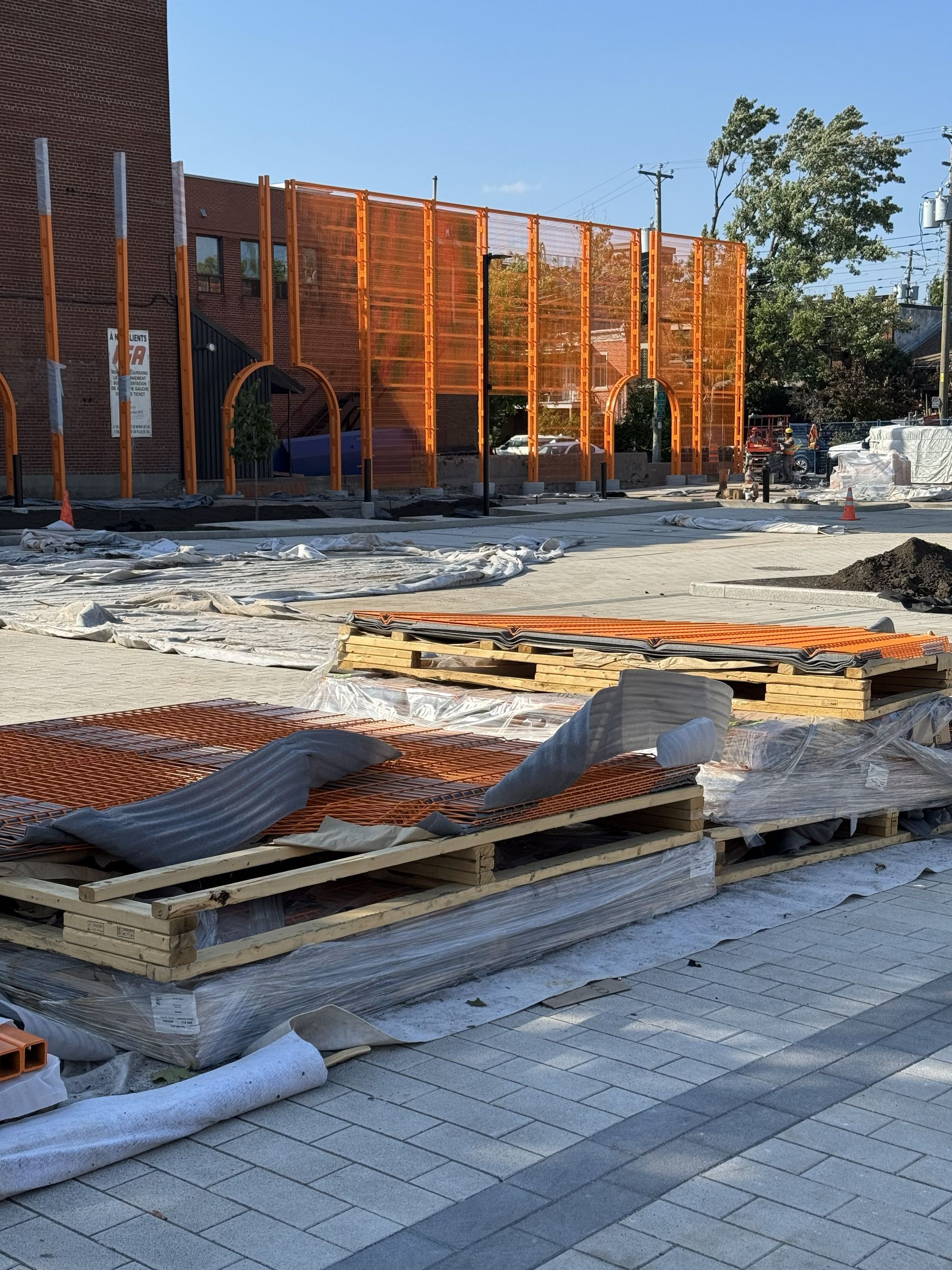 Construction site with orange metal structure, stacked pallets, and building. Paved ground. Sunny day.