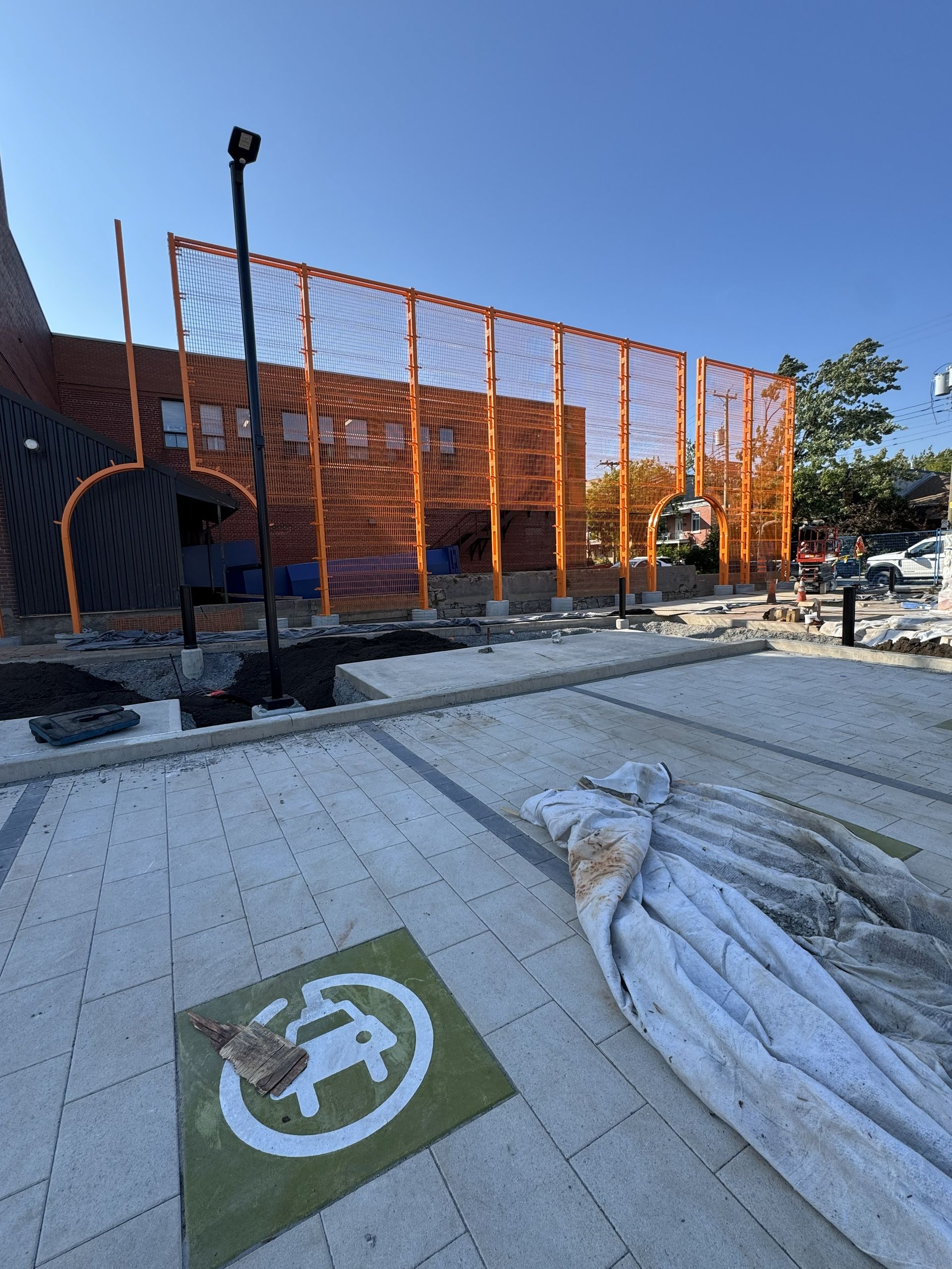 Exterior shot of an orange mesh fence surrounding a parking area. An electric vehicle charging station symbol is visible.