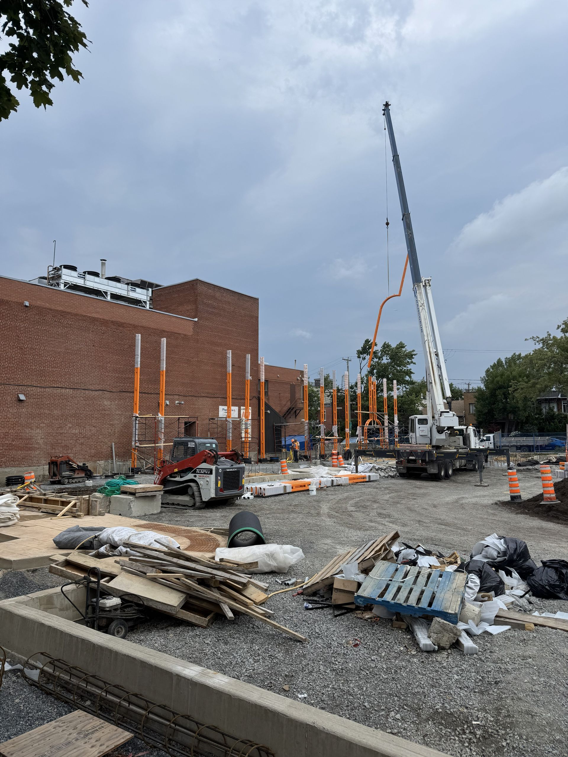 Construction site: crane pouring concrete, brick building, heavy machinery, cloudy sky.