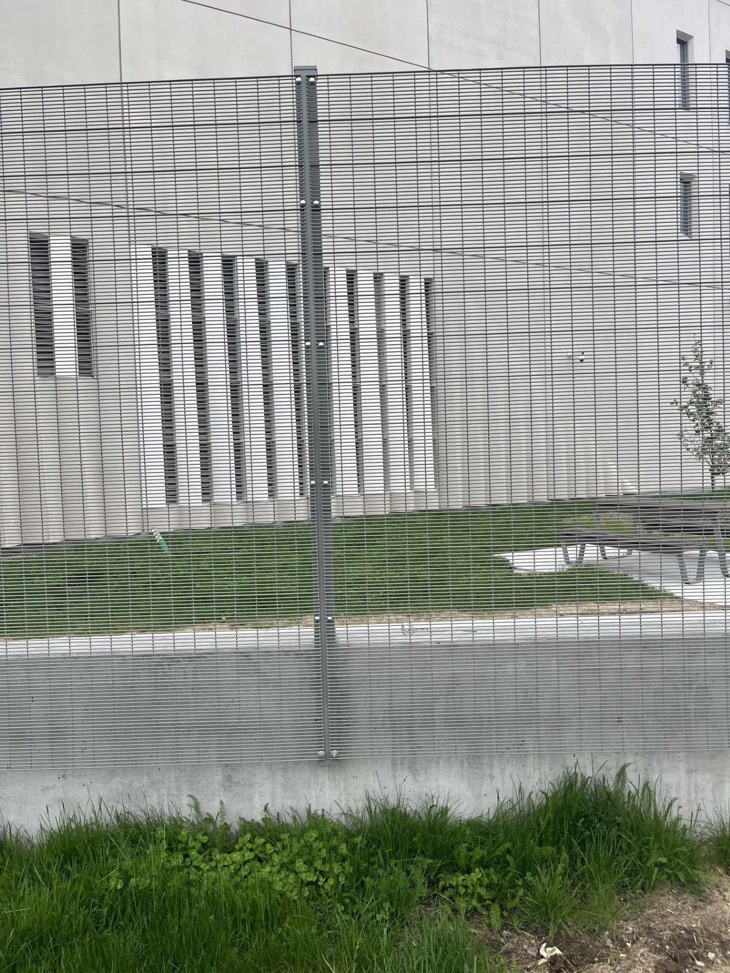 Fence casting a shadow on a concrete wall with windows, grass in foreground.