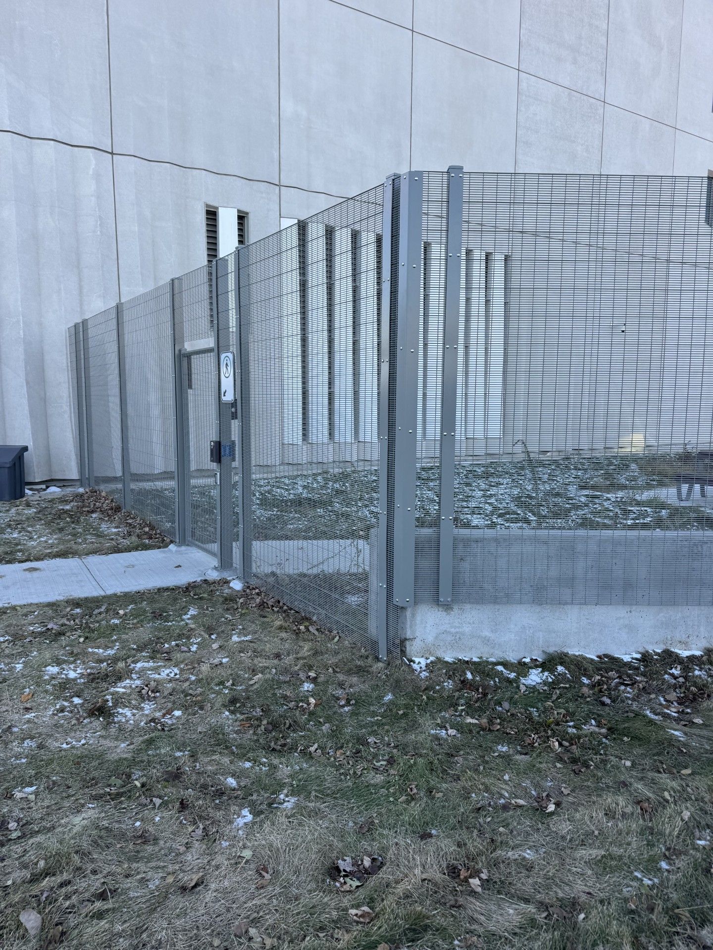 Metal fence with gate, near a building. Ground has grass and patches of snow.