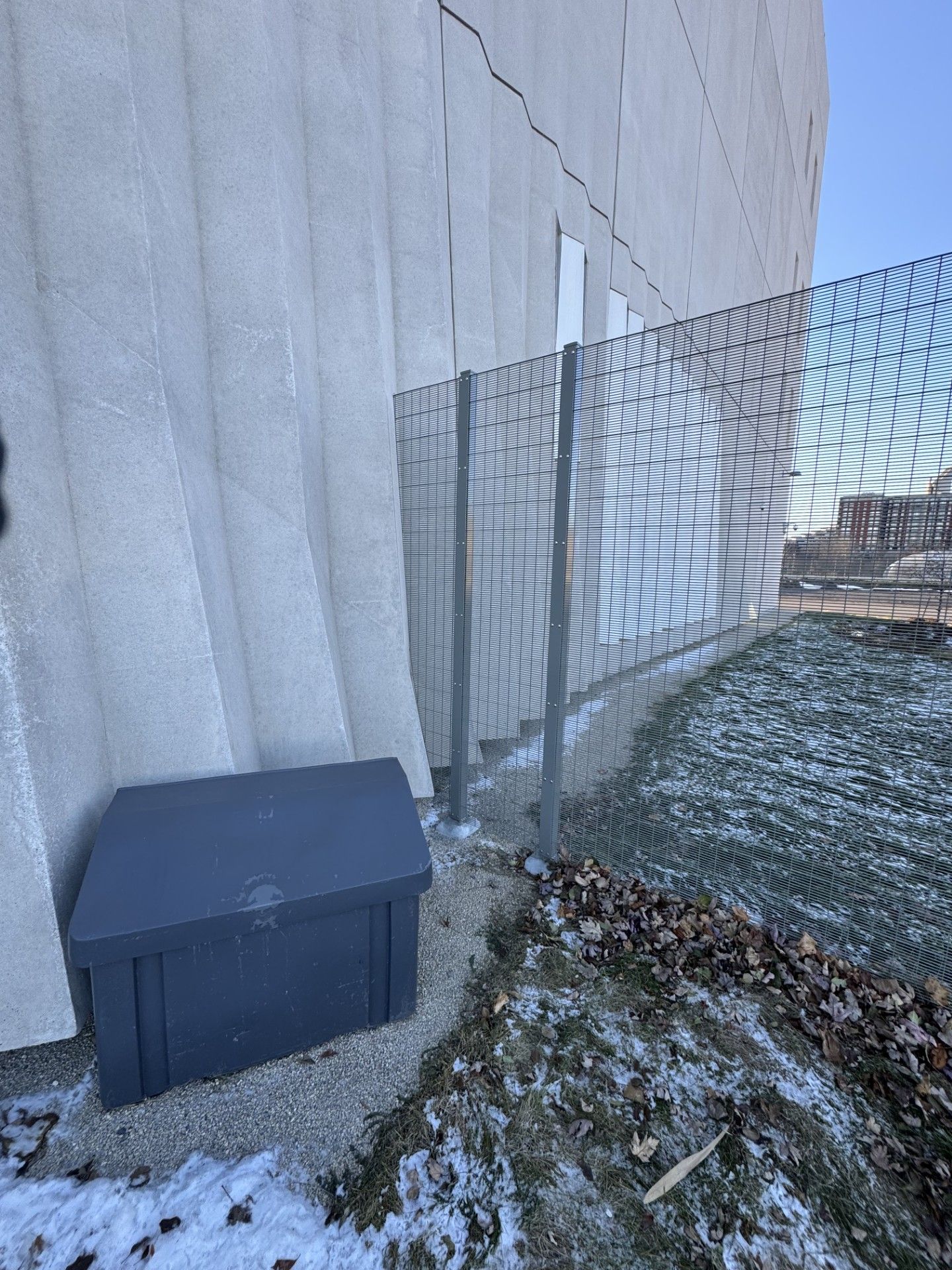 A gray box sits next to a textured building wall and a chain-link fence, with patches of snow.