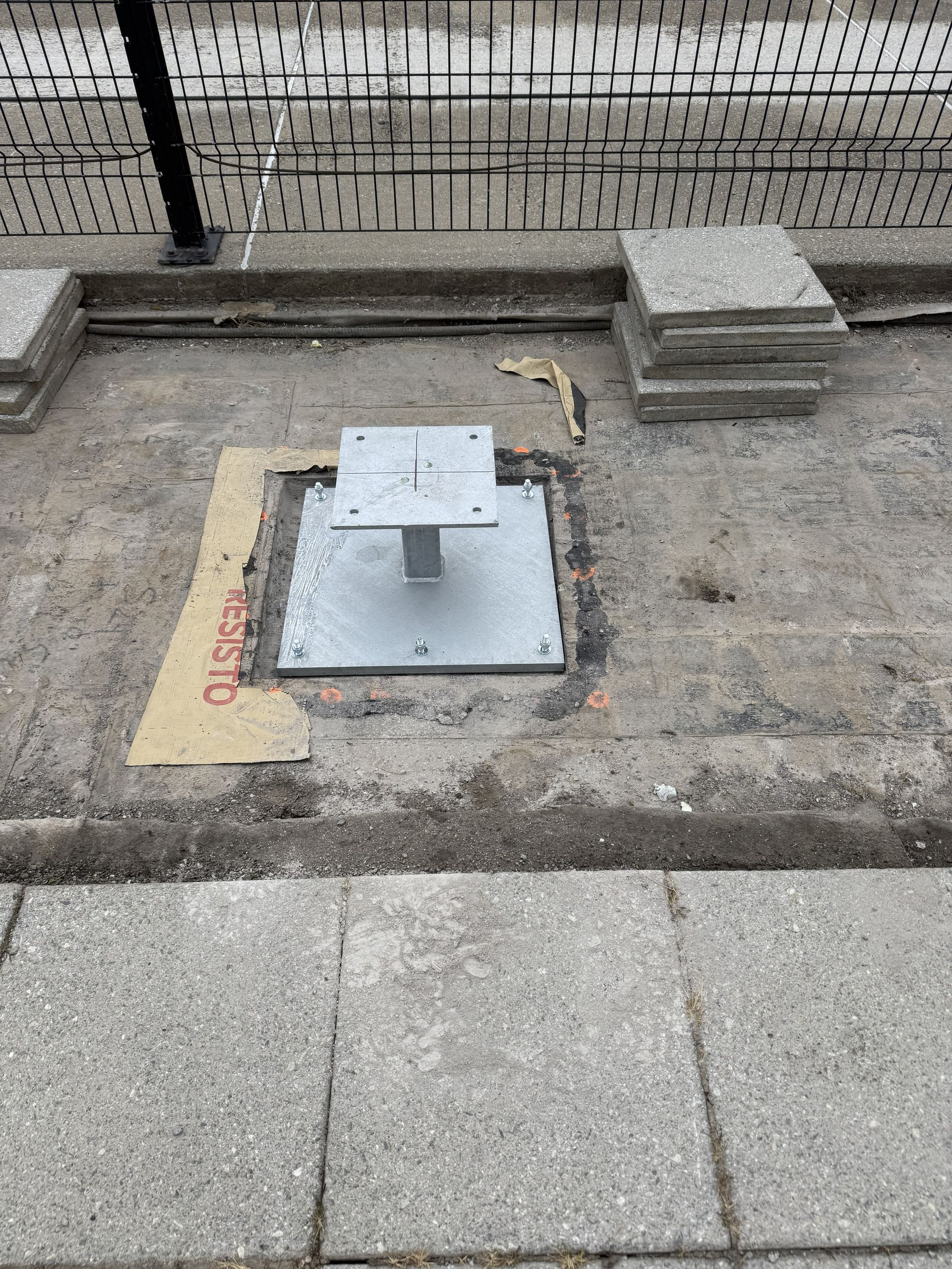 Metal post base installed in a paved area, with nearby concrete pavers and a stack of blocks.