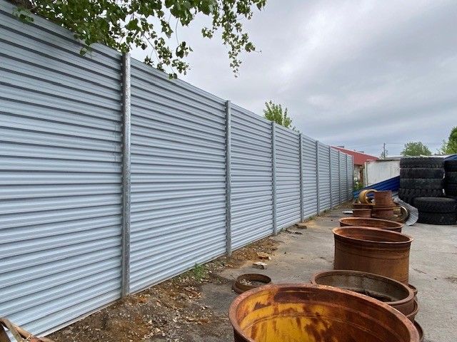 Gray corrugated metal fence bordering an industrial area; rusty barrels and tires are visible.