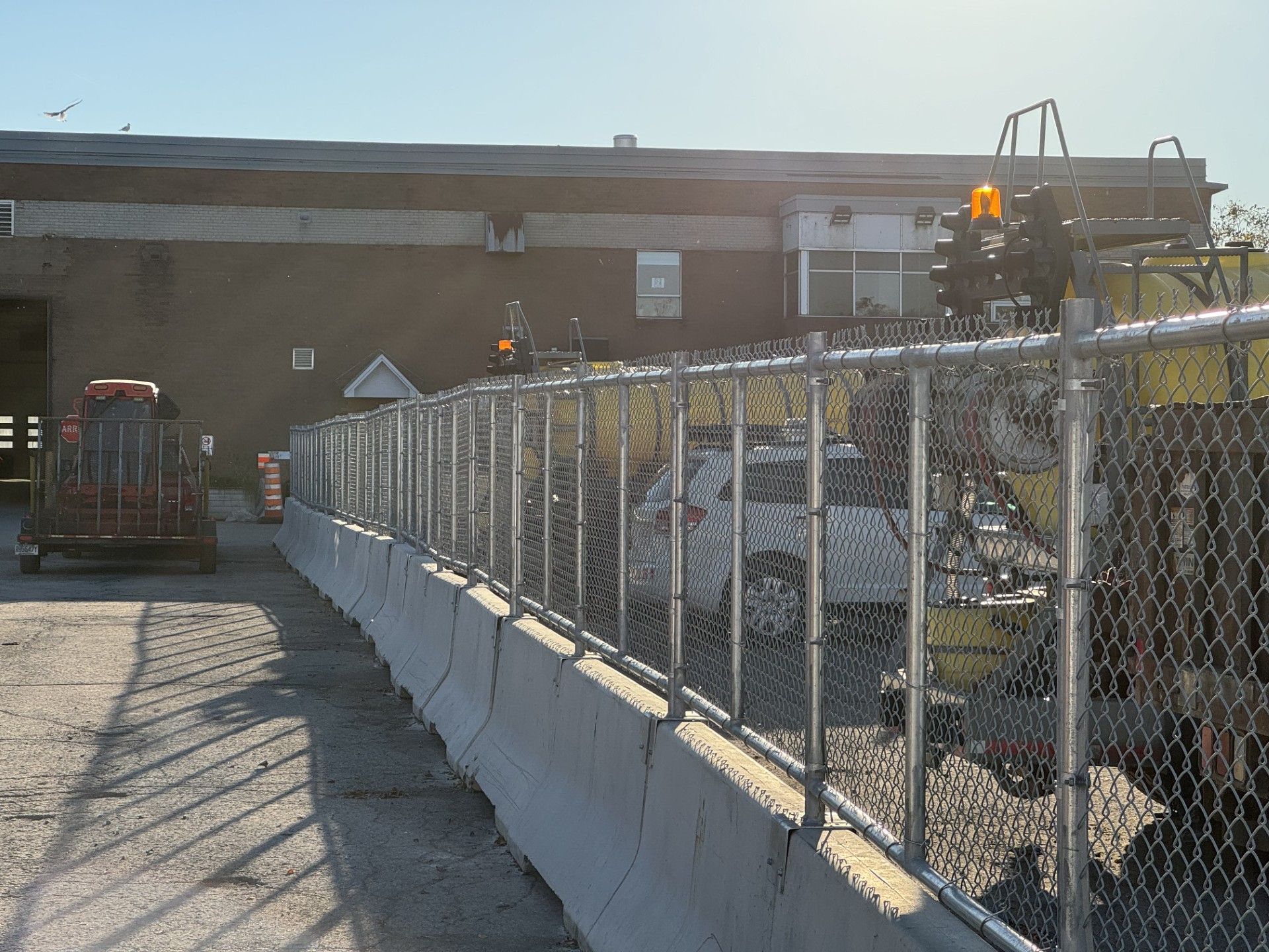 Chain-link fence with concrete barriers along a driveway, with a building in the background. Construction vehicles present.