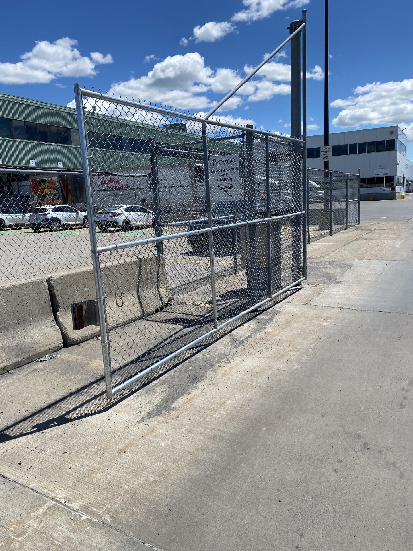 Chain-link fence gate on concrete barrier, commercial building in background, blue sky.