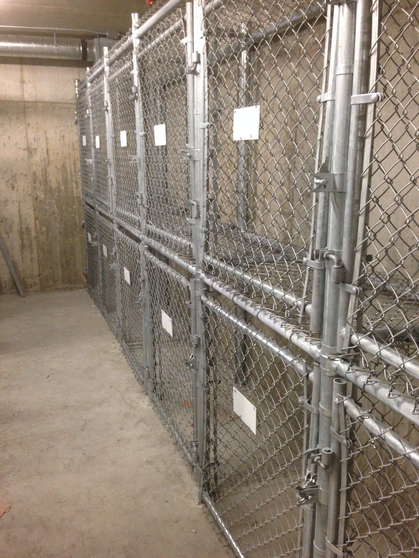Metal storage cages in a concrete room. Chain link fencing with doors and white labels.