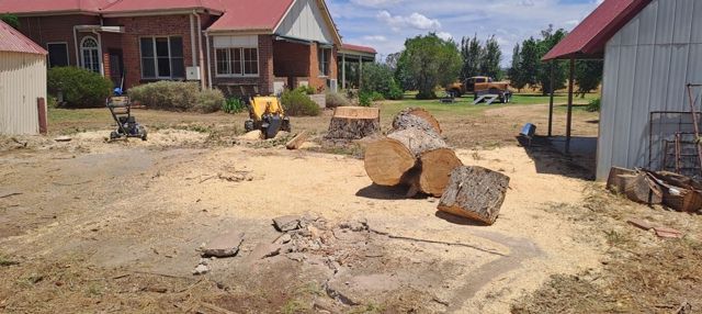 Construction site with a yellow excavator and large cut logs on a dirt lot beside houses