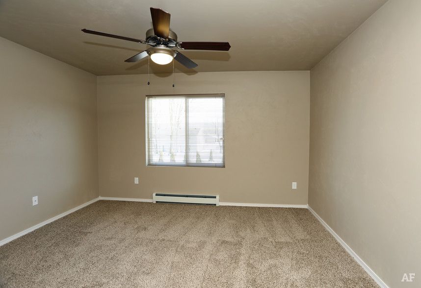 Empty beige bedroom with carpet, ceiling fan, and a window above a baseboard heater