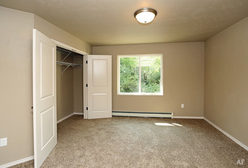 Empty beige bedroom with carpet, open closet, white door, and window overlooking greenery.