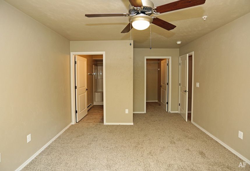 Empty beige bedroom with carpet, ceiling fan, and open doors to a closet and bathroom