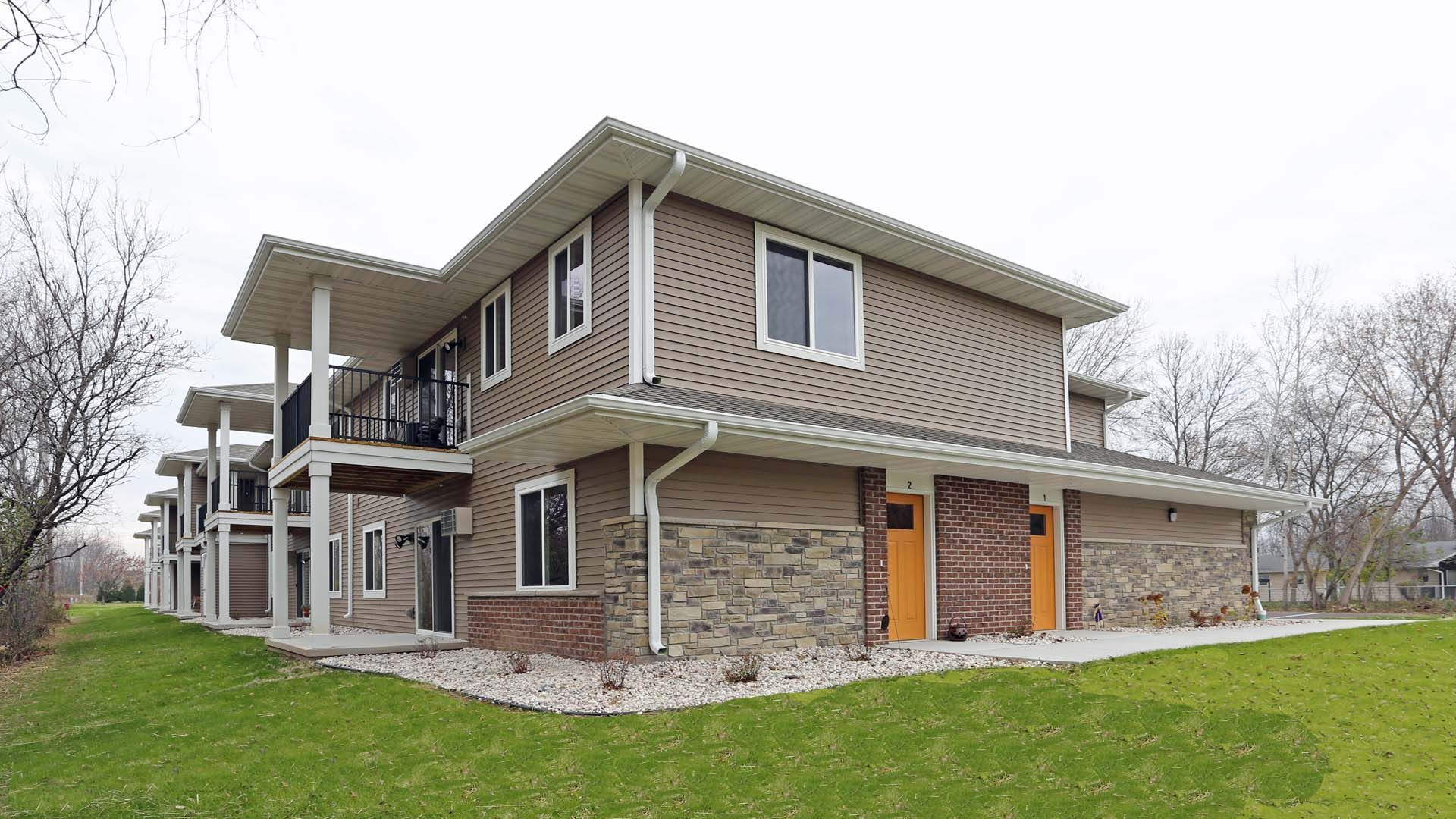 Two-story house with stone and tan siding, seen on a grassy lawn under an overcast sky.