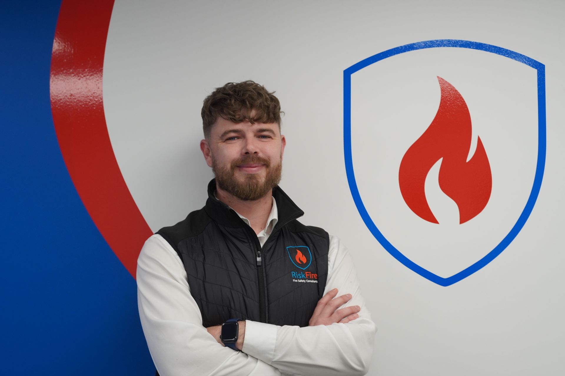 Man in vest with arms crossed, standing near wall with fire safety logo.