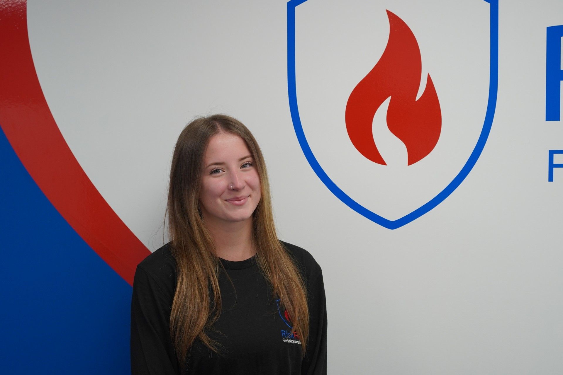 Woman stands in front of a wall with a shield logo depicting a red flame, with blue and red details.