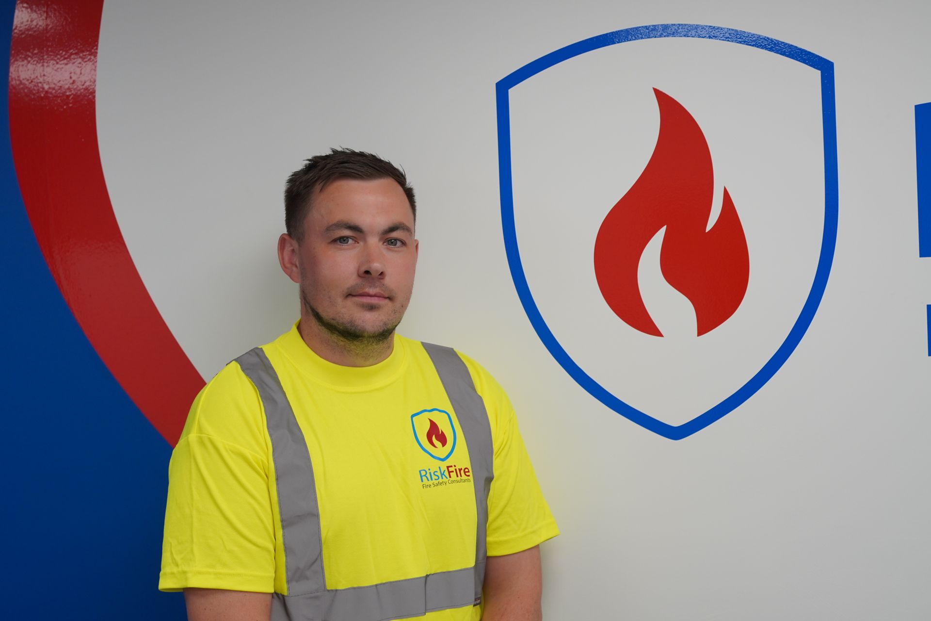 Man in yellow shirt with arm tattoos, standing in front of a company logo with a flame icon.