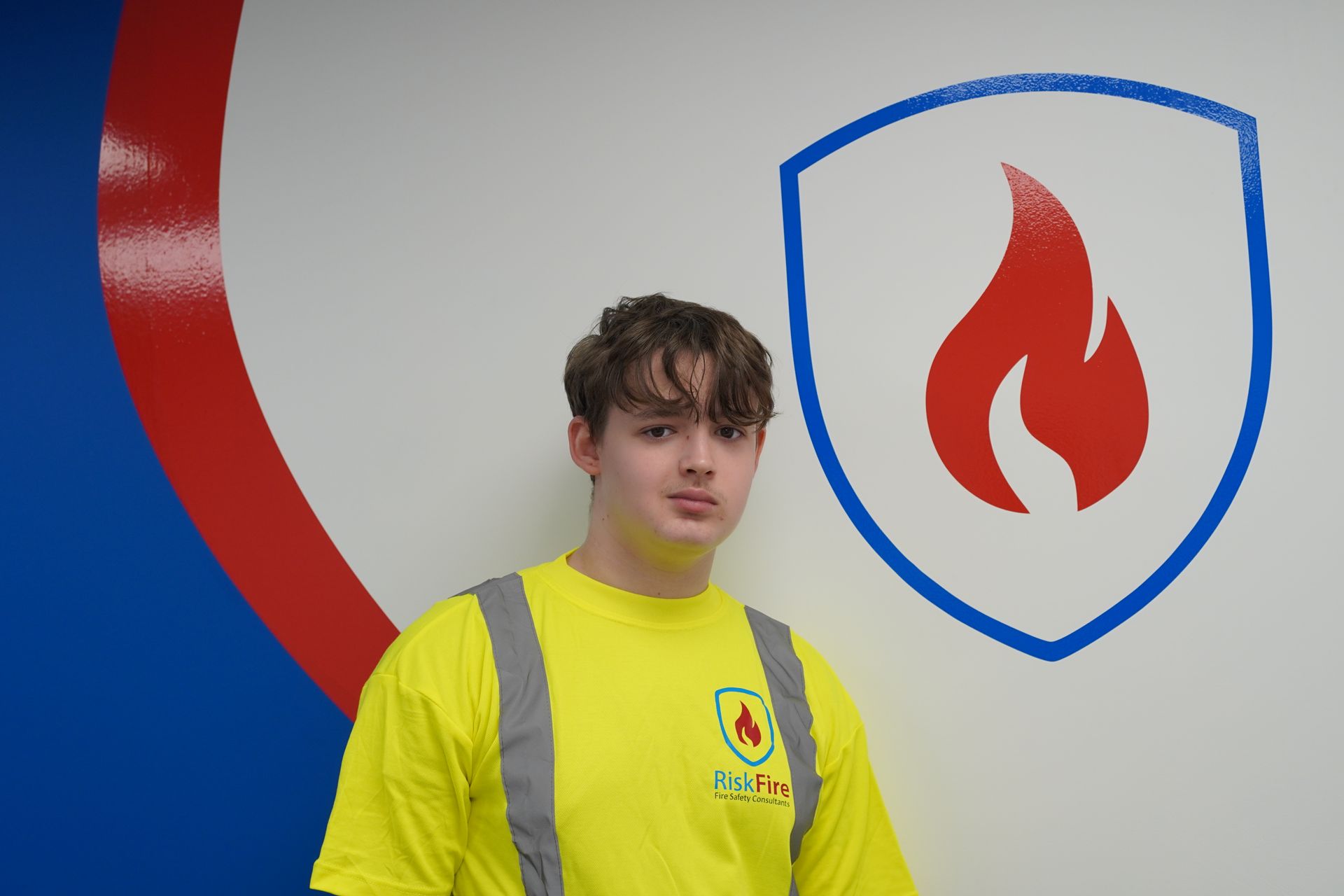 Young person in yellow safety vest stands in front of a wall with a fire department logo.