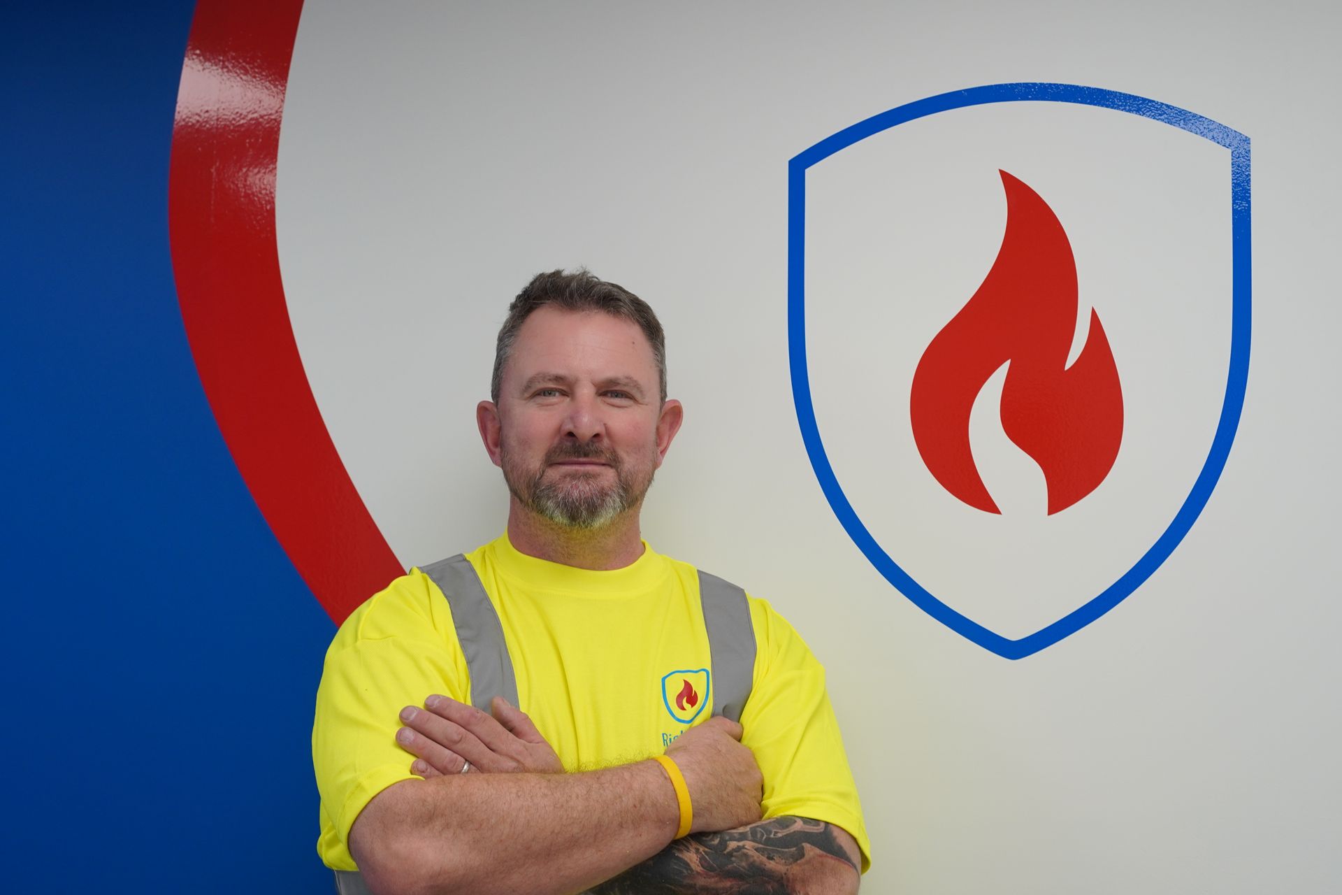 Man in yellow safety shirt, arms crossed, stands before a shield logo with a red flame.