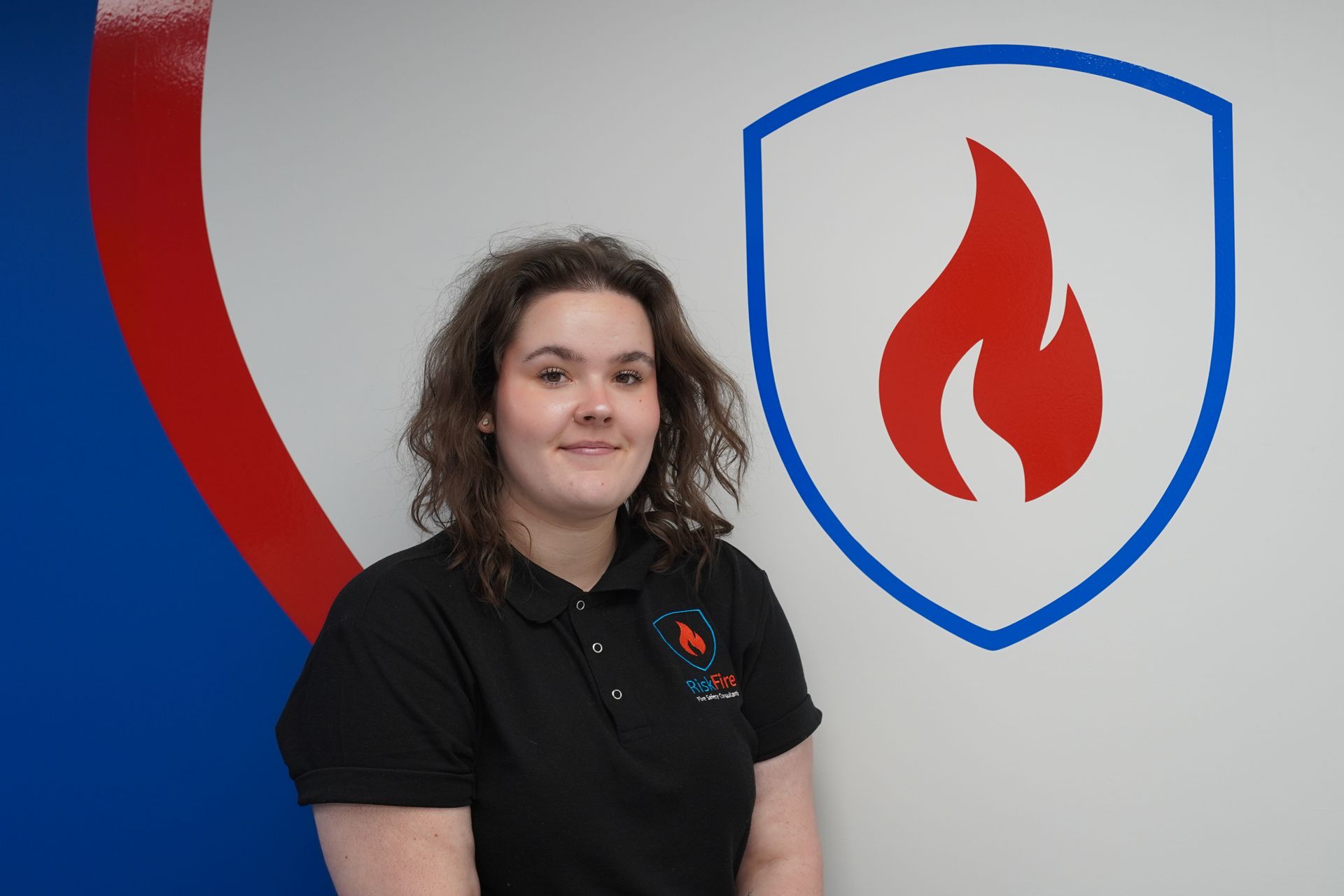Woman in black shirt smiles next to logo of a red flame within a blue shield on a white wall.