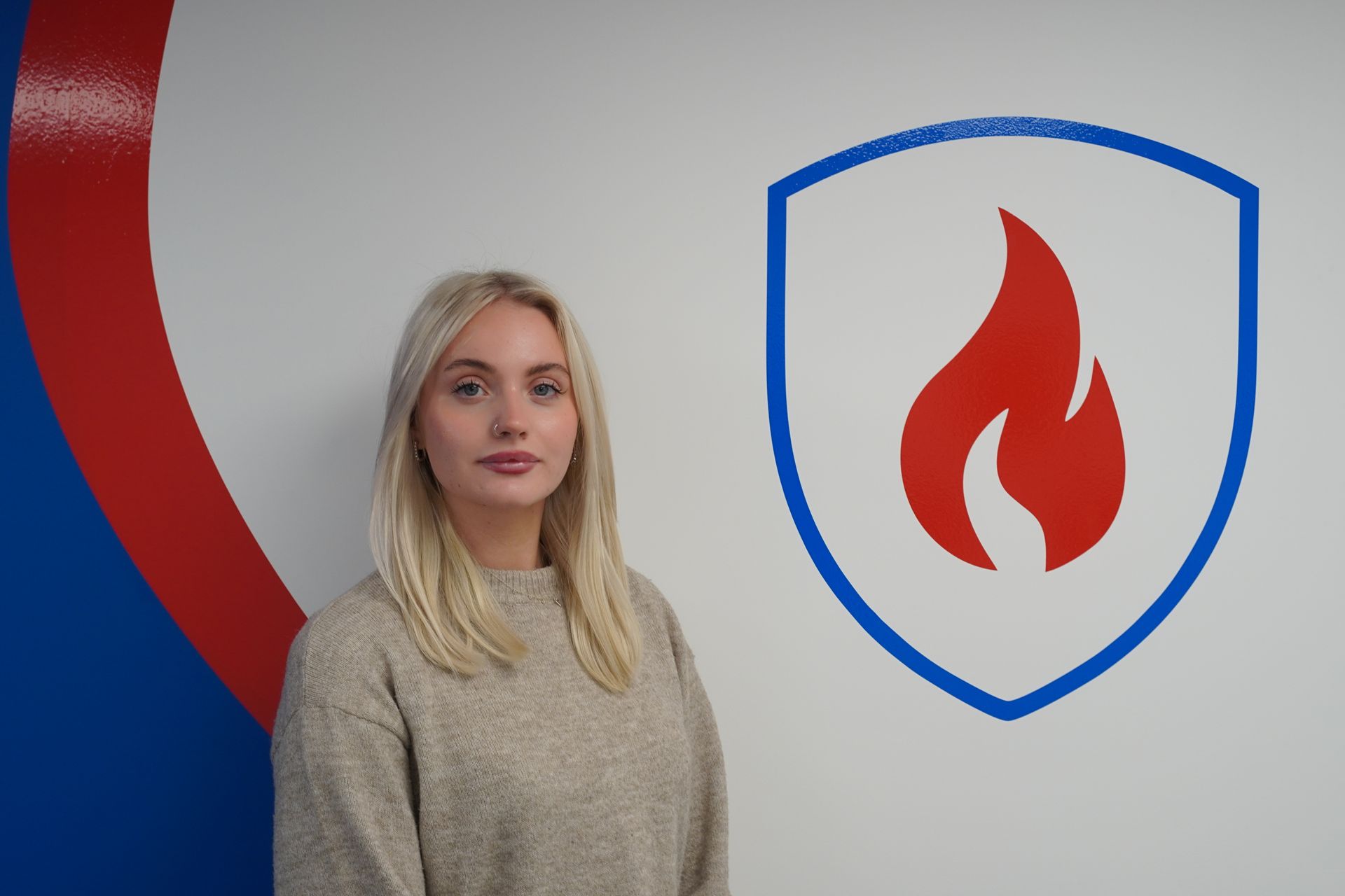 Woman stands in front of a wall with a shield logo depicting a red flame, with blue and red details.