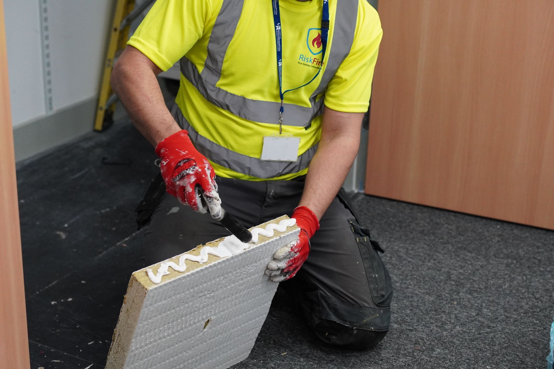 Person kneeling, applying adhesive to a panel, wearing gloves, a yellow vest, and safety glasses indoors.