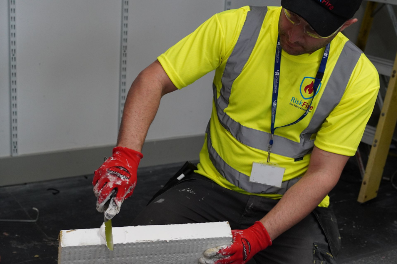 Construction worker in safety vest using a knife to cut a light-colored brick.