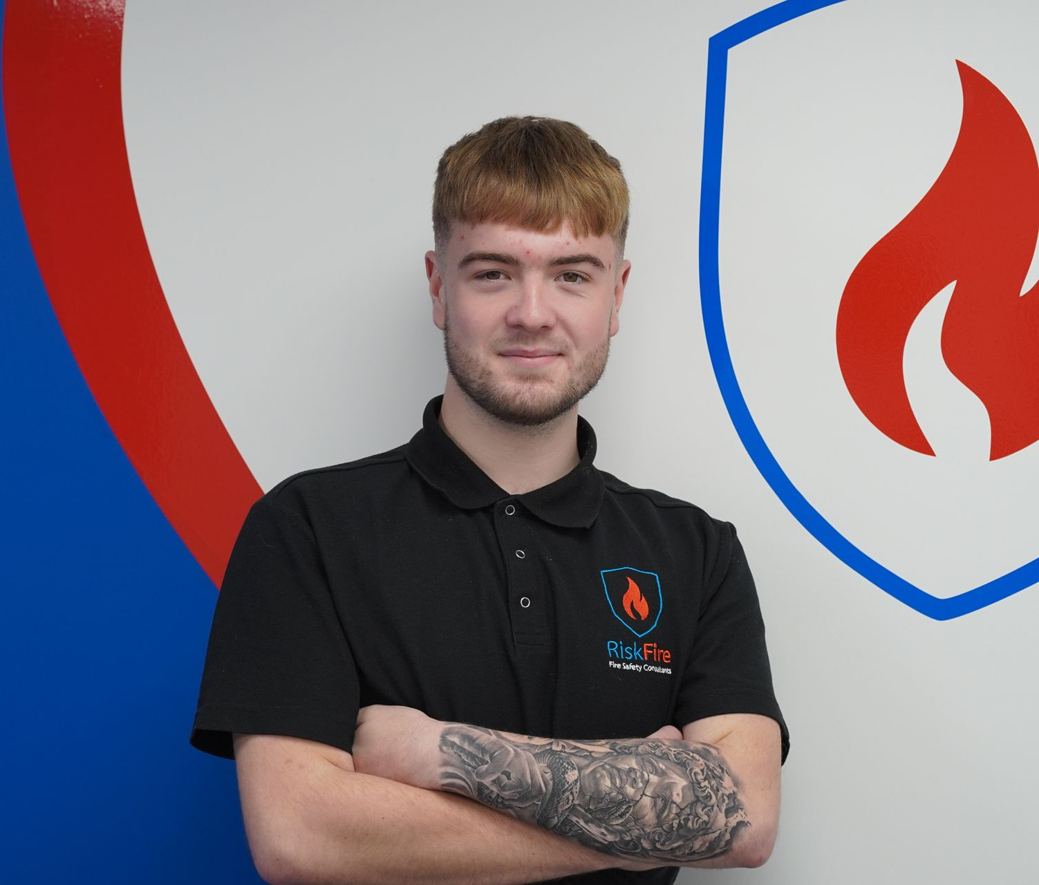 Man in black polo shirt with crossed arms, smiling, in front of a blue and red shield graphic.