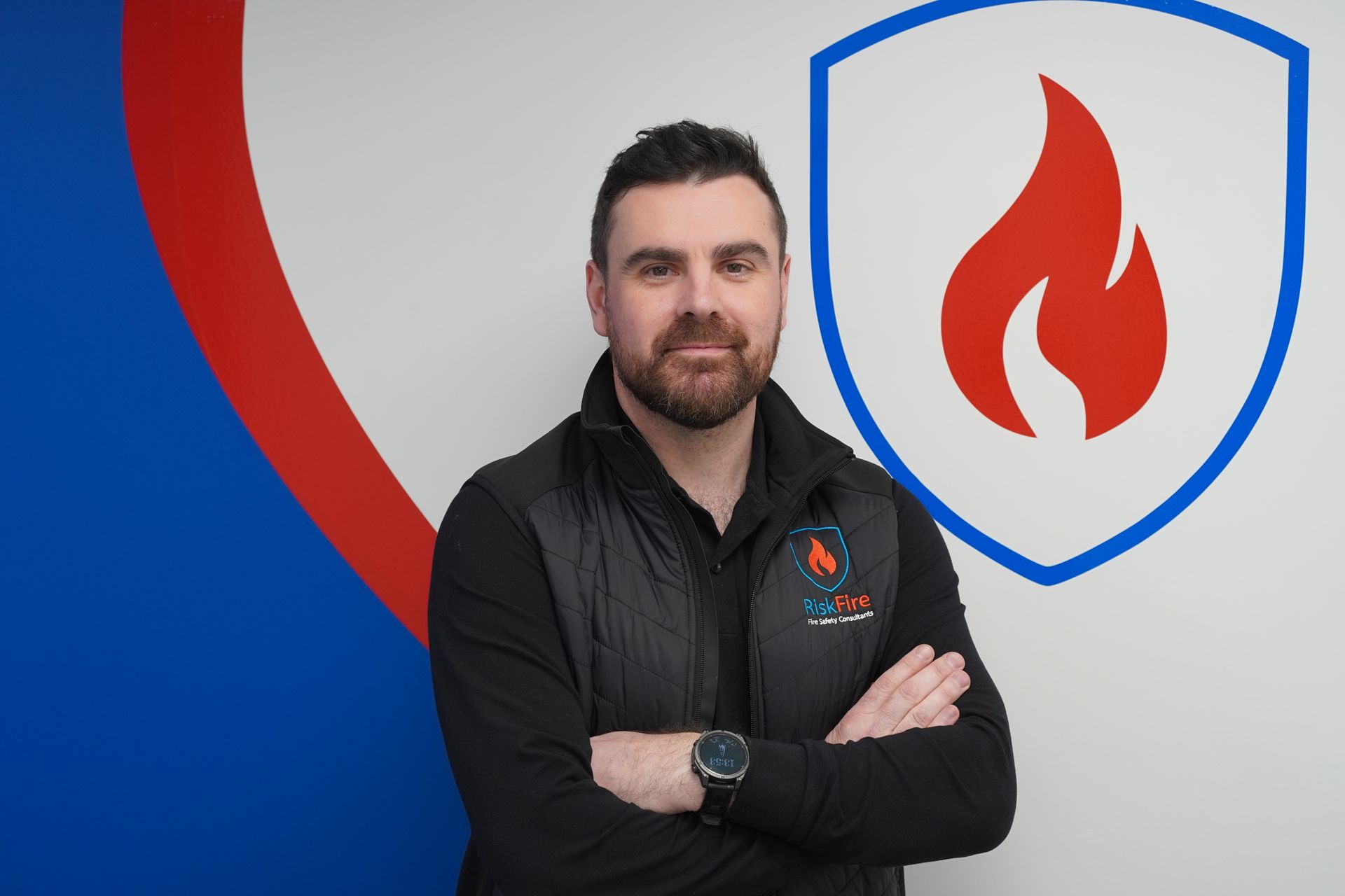 Man with crossed arms in front of a blue and red shield logo with a flame.