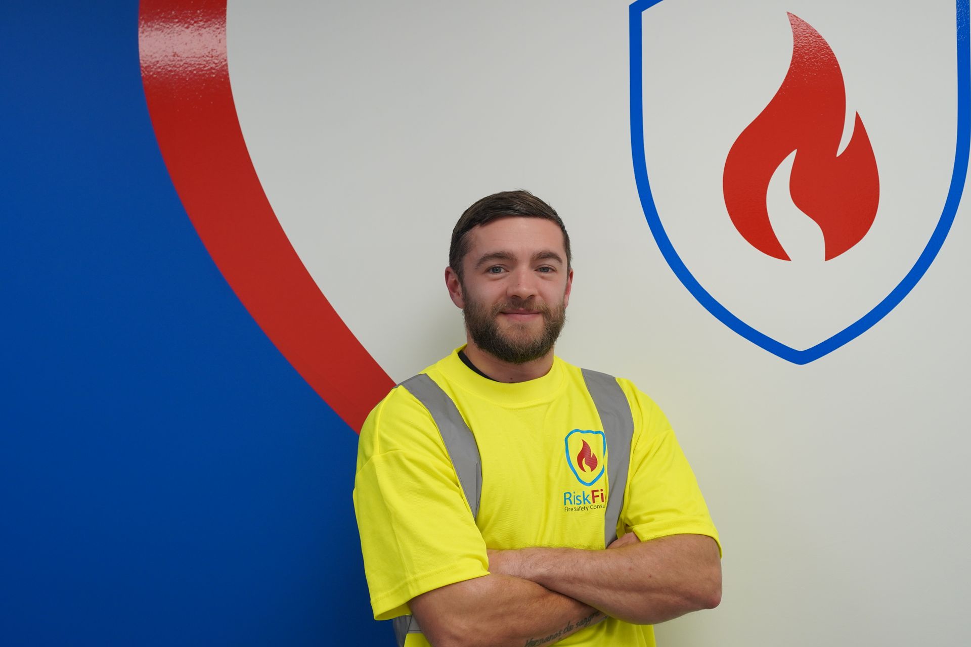 Man in yellow safety vest with crossed arms in front of blue and red logo.