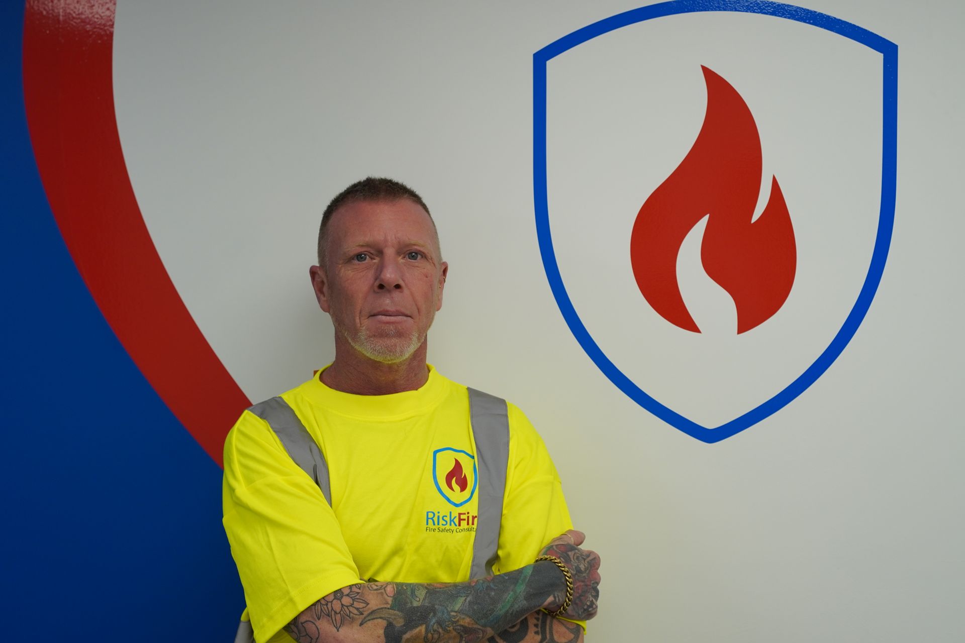 Man in yellow shirt with arm tattoos, standing in front of a company logo with a flame icon.