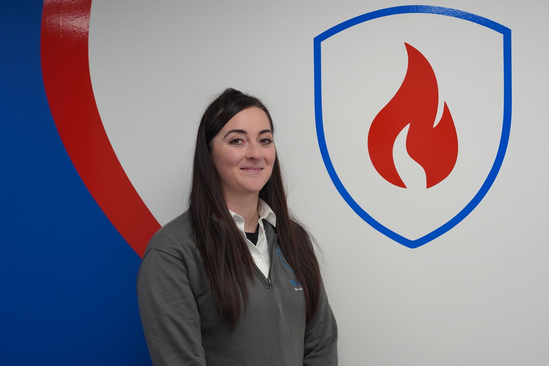 Woman in gray sweater smiling, standing in front of a wall with a red and blue shield logo with a flame.