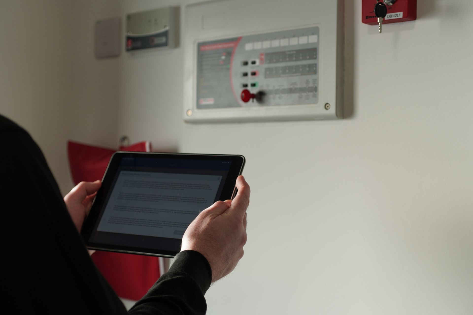 Person holding tablet, facing a fire alarm panel on a white wall.